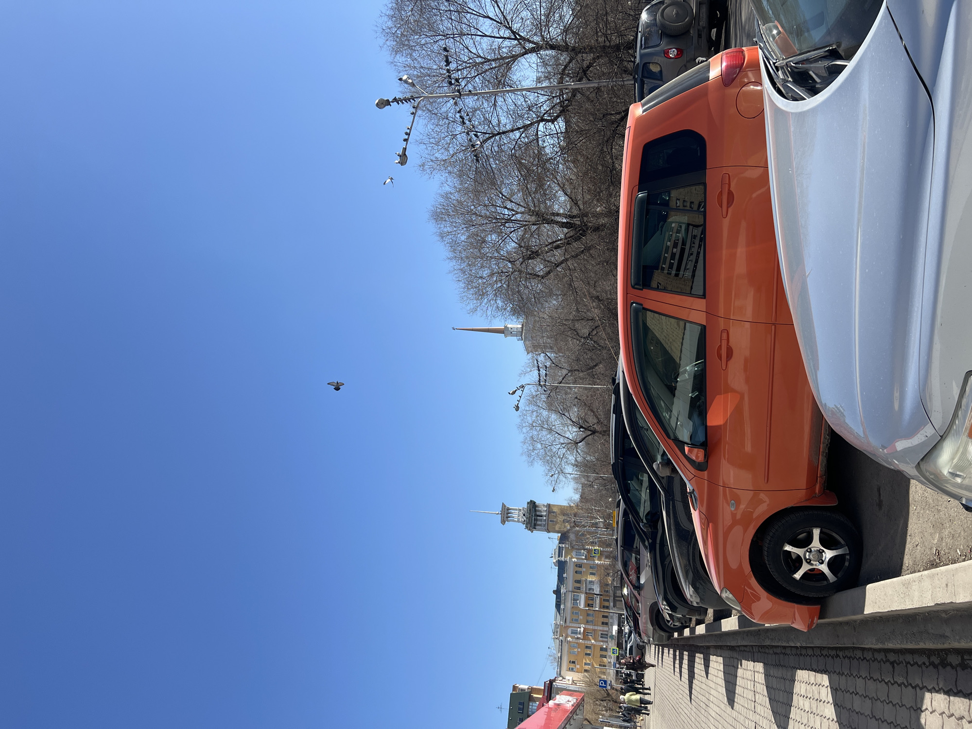 The image captures a street scene in Krasnoyarsk, Russia, characterized by parked cars lining the right side of the frame. The cars are of various colors, including orange, silver, and black. A sidewalk is present to the right of the cars, and pedestrians are visible in the background, suggesting street activity.
On the left side of the frame, the sky is a clear, bright blue. In the background, a street with bare trees and buildings can be seen. A distinct tower is noticeable amongst the buildings. Birds are visible in the sky. The overall setting suggests an urban environment on a clear, sunny day.