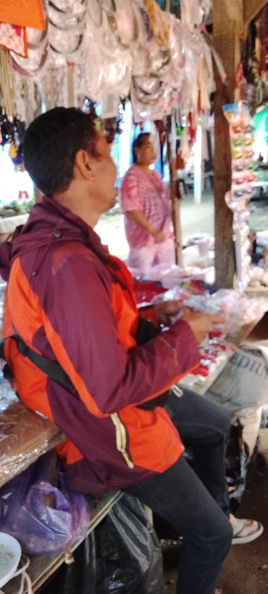 A low-angle, outdoor, medium shot captures two individuals in what appears to be a marketplace stall in Bahway, Indonesia. In the foreground, a person wearing a purple and orange hooded jacket with a yellow zipper and dark pants is seated and holding items in their hands, partially obscured from view. Their back is turned towards the camera, and they are looking to the right. Behind them and slightly to the right, another person wearing a pink tie-dye shirt and white pants is standing, also looking to the right. The background is filled with an array of merchandise displayed on racks and tables, including what appear to be colorful accessories, some packaged in plastic. The environment suggests a bustling market with a variety of goods for sale.