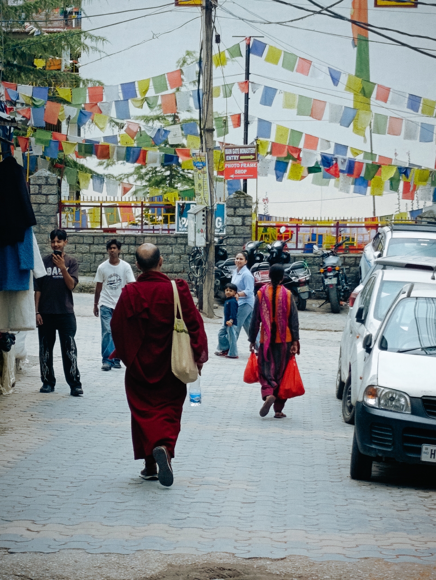 This is a photograph taken on a street in Manali, India, likely near a monastery given the prayer flags overhead. The street is paved with interlocking gray stones and is lined with buildings and a stone wall. Various people are walking in the street. In the foreground, a person in a dark red robe walks away from the camera, carrying a beige bag over their shoulder and a clear plastic bottle in their hand. To the left, a young man in a dark t-shirt and pants is looking at his phone. Behind him, another young man in a white t-shirt and jeans walks past. Further down the street, a woman in a blue shirt and jeans walks with a young child. A woman in a colorful tunic and pants carries two bright red shopping bags. Several motorcycles and white vehicles are parked on the street. Prayer flags in various colors are strung across the street and between poles.