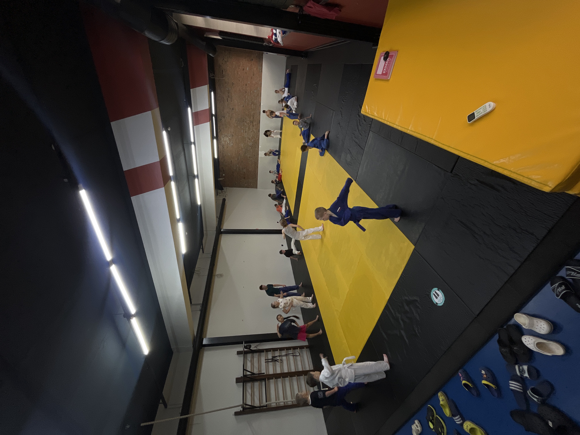 The image depicts an indoor judo dojo where children and adults are participating in a training session. The dojo features a large yellow mat surrounded by black mats. A wall-mounted climbing apparatus is visible on the left. The room is well-lit by fluorescent lighting. Some individuals are wearing judogi (judo uniforms), while others are in athletic wear. The individuals appear to be engaged in warm-up exercises or practice. The background shows a brick wall. Scattered on the floor near the mats are various pairs of footwear, including sandals and flip-flops. The scene is consistent with a martial arts training facility. This location is identified as a judo club in Krasnoyarsk, Russia.