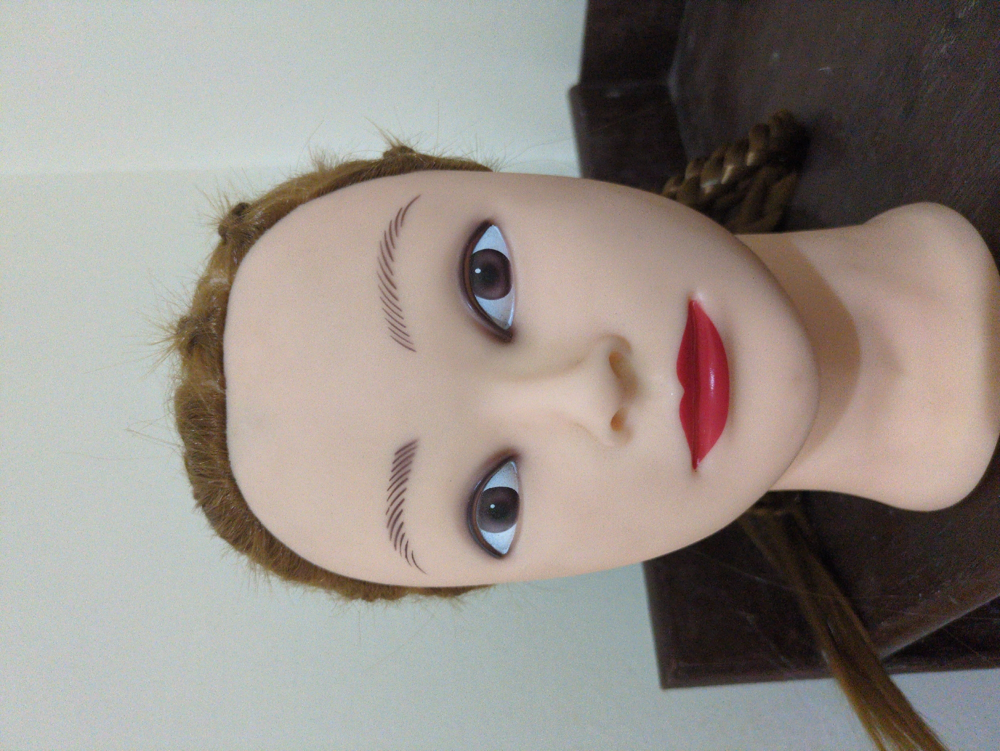 The image shows a close-up, head-on view of a mannequin's head. The mannequin has light tan skin, brown eyes with defined eyelashes and painted eyebrows. Its lips are painted bright red. The mannequin has light brown hair, which is partially styled with some strands pulled back and a braid visible on the right side of its head. Some loose strands of hair extend from the head and to the left. The mannequin is positioned in front of a neutral, light-colored wall. The bottom portion of the image shows a dark brown surface, possibly a table or stand, on which the mannequin is resting. There are no discernible geographical markers to indicate it is located in Haini, India.