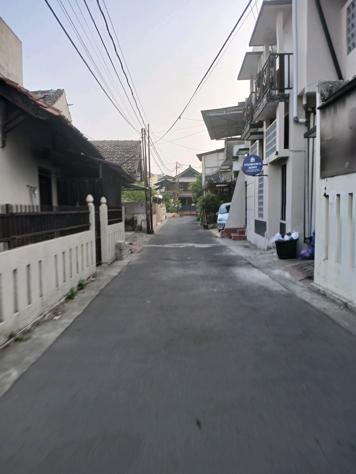 The image shows a narrow asphalt street in Depok, Indonesia, lined with residential buildings and a fence. The sky is overcast. On the left side of the street, there is a white fence with vertical slats and a dark metal gate. Behind the fence, a building with a dark tiled roof and a light-colored wall is visible. On the right side, a multi-story building with balconies and white walls stands. A circular blue sign with white text that reads "PANDAWA KOST" is attached to the building. A white car is parked on the side of the road, and a black trash bin with trash bags is near the building entrance. Overhead, numerous electrical wires are suspended across the street. The overall impression is of a residential area in a city.