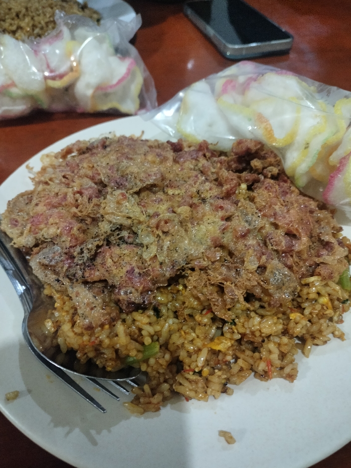 The image displays a close-up, eye-level view of a plate of Indonesian fried rice, known as Nasi Goreng, served on a white circular plate. The fried rice itself is a brownish-orange color, with visible specks of green vegetables and possibly other ingredients.
The prominent feature of the dish is a large, crispy fried egg, likely a "dadar" style omelet, which covers a significant portion of the rice. It exhibits a dark golden-brown hue and a distinctive lacy, crunchy texture. A silver-colored fork and spoon are placed on the left side of the plate, with their heads resting within the fried rice.
In the blurred background, two clear plastic bags containing kerupuk (crackers) are visible. These crackers are predominantly white with colorful edges in pink, yellow, and green. To the right, a dark-colored smartphone lies face down on a dark brown table, reflecting some ambient light. Another plate with food, possibly more fried rice, is partially visible in the upper left background, out of focus.
The scene suggests a casual dining setting, possibly indoors during the evening or night, given the somewhat dim lighting. The overall impression is of a traditional Indonesian meal. No discernible text is visible in the image.