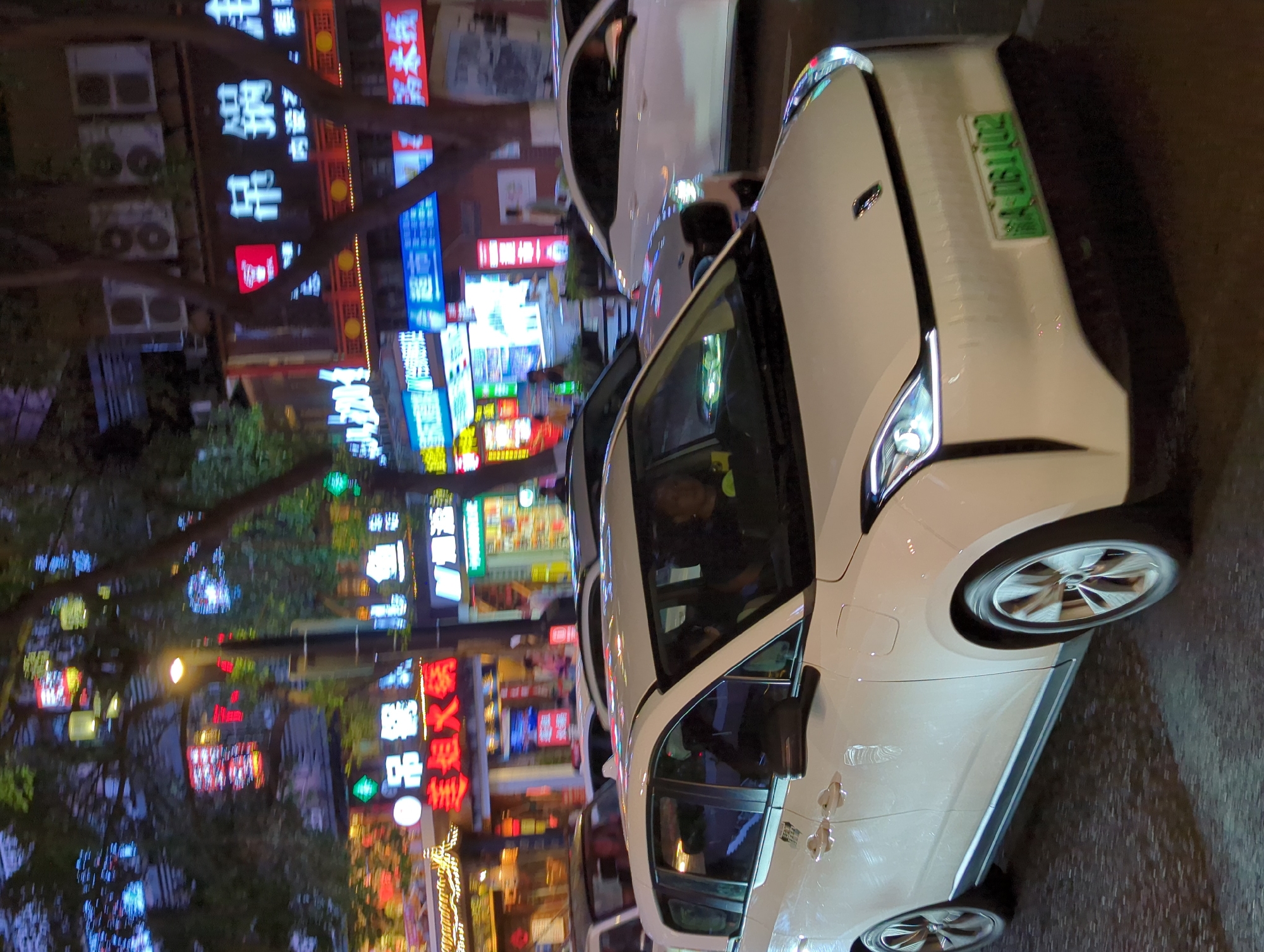 A white electric vehicle with a green license plate displaying "A 061102" is captured in motion at night on a street in Yuzhong District, China. The car is the primary subject, positioned in the foreground and taking up a significant portion of the frame. Its headlights are illuminated, casting light on the wet asphalt road. The vehicle appears to be part of a convoy of cars, with other vehicles visible in the background, some of which are also white. The environment is characteristic of a bustling city street at night, with numerous neon signs and illuminated storefronts lining the buildings. These signs, in Chinese characters, suggest commercial activity, likely restaurants and shops, as indicated by text such as "美姐火锅" (Mei Jie Hot Pot) and other unintelligible characters. Trees with visible foliage are interspersed with the buildings and signs, their branches creating a canopy over the street. The lighting is predominantly artificial, with vibrant colors from the signs reflecting on the wet road and vehicles. One person is visible inside the electric vehicle, behind the steering wheel, appearing to be the driver. The overall impression is a dynamic urban nighttime scene, with traffic and commercial lights creating a lively atmosphere.