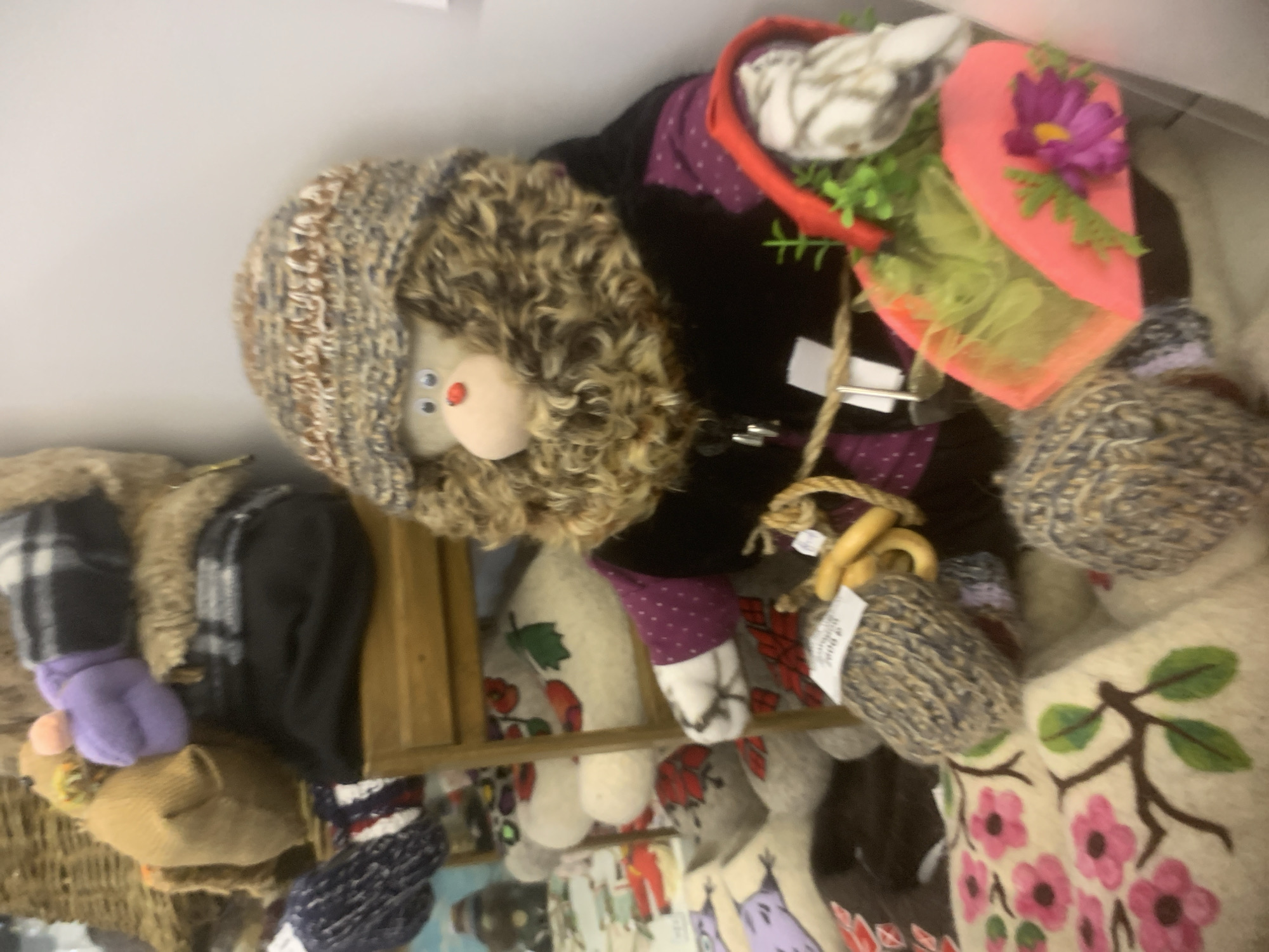 The image appears to be a cluttered display of handcrafted dolls and related items. The location cannot be confirmed as Izhevsk, Russia, from the image alone.
**Objects:** Several dolls with varied designs and fabrics are visible. One doll has a light-colored face, curly brown hair, a knitted cap, and a red nose. Another doll wears a pink hat with a purple flower. A black jacket or coat with purple accents is also present. Other fabric items, including knitted balls and decorative cloths with floral designs, are mixed in. A wooden shelf is also visible.
**Environment:** The scene suggests an indoor setting, possibly a shop or a market stall, given the density of items displayed. The lighting appears to be diffused and somewhat dim.
**Actions/Interactions:** The items are arranged in a display, suggesting they are for sale. There are no people visible in the image and no explicit interactions taking place.