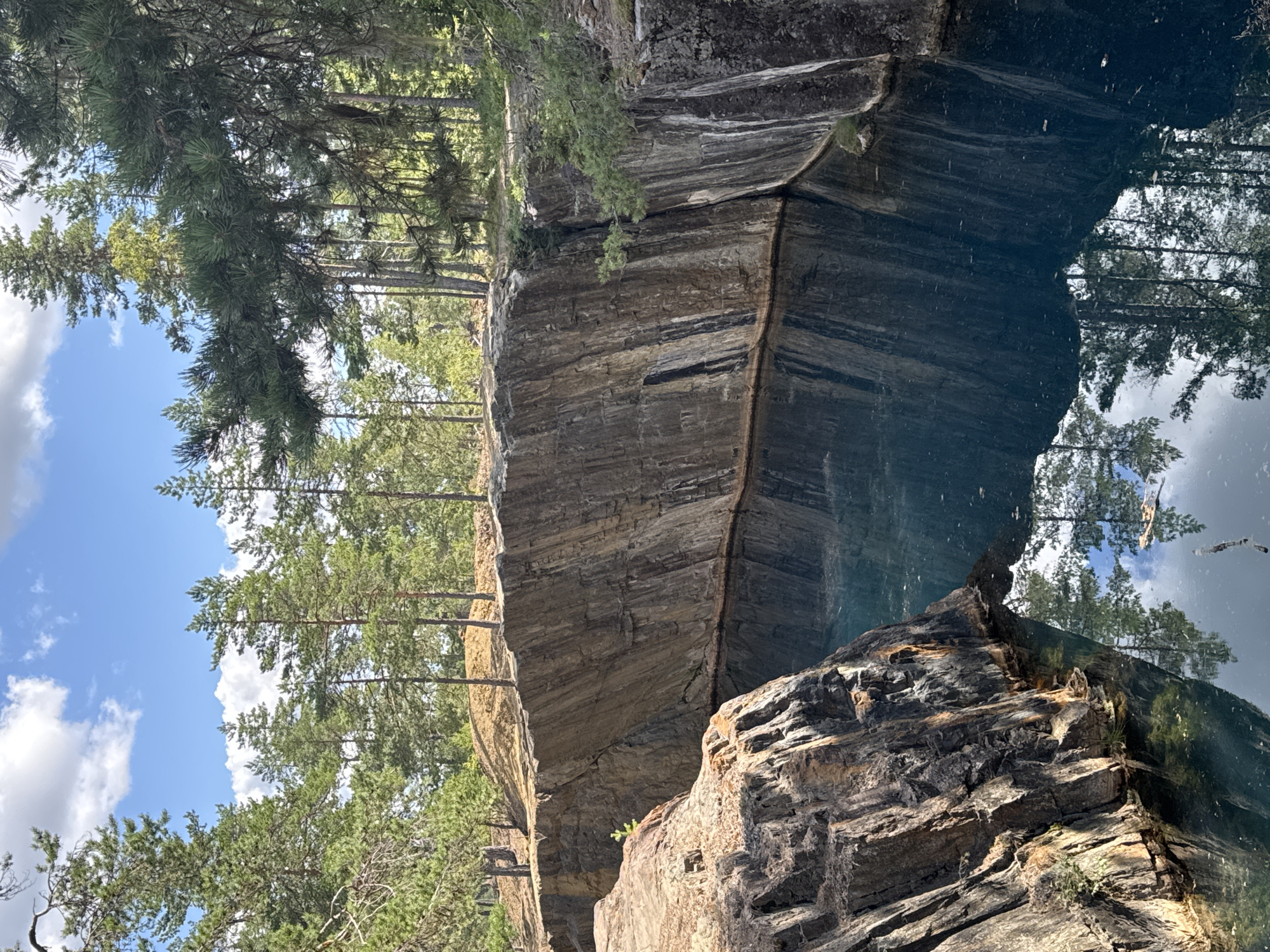 The image shows a quarry in Säter, Sweden, with steep, rocky walls surrounding a dark, reflective body of water. Pine trees line the top of the quarry, with their reflections visible in the water. The sky above is blue with scattered white clouds. The rock faces are textured and show layers of different colors, primarily brown and gray, with some white mineral deposits. The water is a deep blue-green and appears still, mirroring the surrounding trees and sky. In the foreground, to the left, a prominent rock formation juts out, also showing textured, layered surfaces. There are no people or animals visible in the image. The overall impression is one of a natural, somewhat rugged landscape.