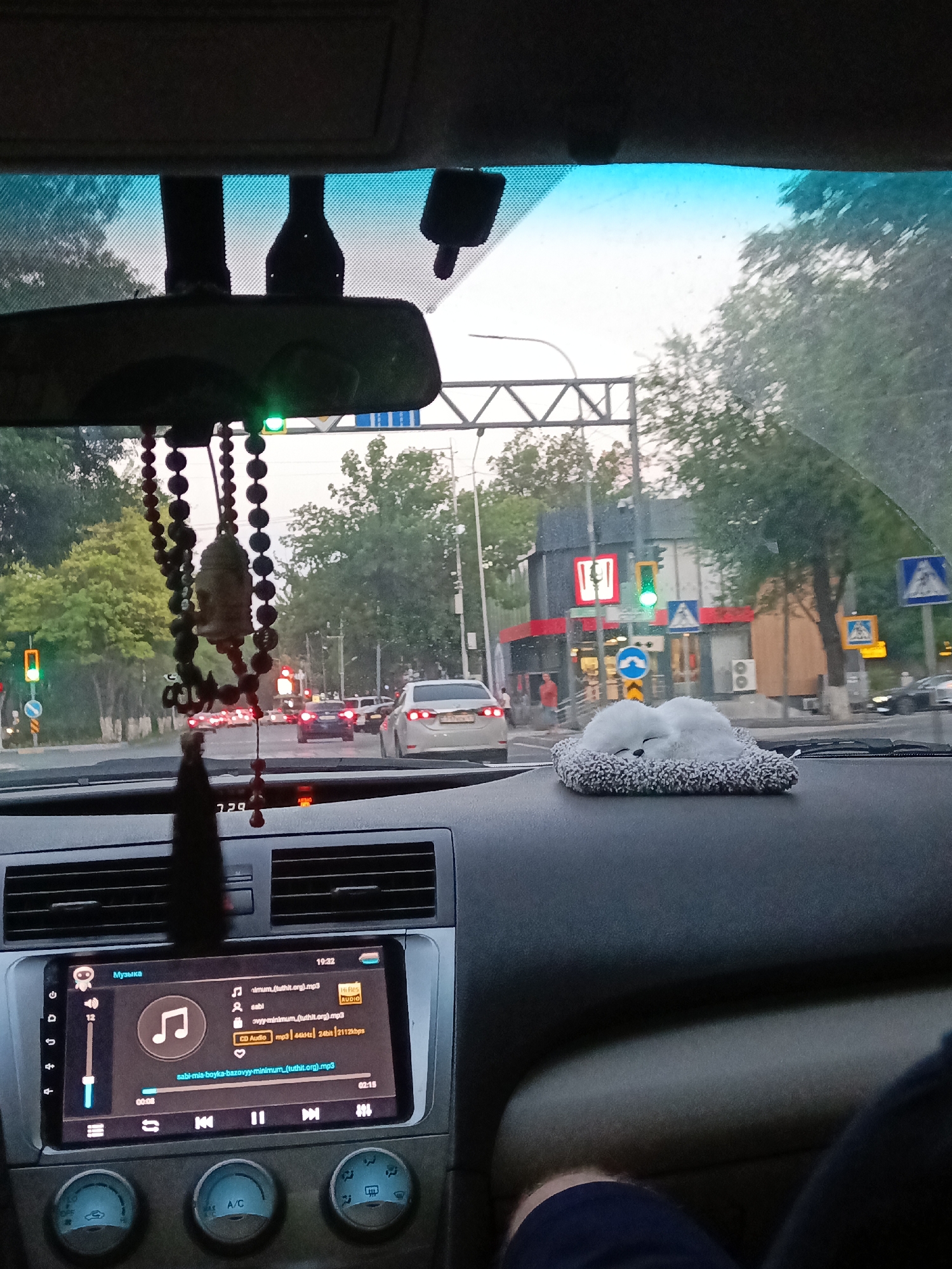 The image is taken from the interior of a car, looking forward through the windshield. A car's rearview mirror is visible at the top, with a green traffic light illuminated above it. Below the mirror, a beaded string with a Buddha-like pendant hangs down. The dashboard of the car is in the foreground, featuring an infotainment screen displaying music playback with the title "minum.tuthit.org).mp3". To the right of the infotainment screen, a fluffy white toy resembling a sleeping animal is placed on the dashboard.
Outside the car, a street scene is visible. Traffic lights show green and orange, indicating an intersection. Several cars are on the road, some stopped at the intersection. Buildings line the street, including one with a prominent red and white logo that appears to be KFC. Pedestrian crossing signs are also visible. The trees along the street suggest an urban environment. Based on the visual cues, particularly the presence of KFC and the general urban landscape, this scene is consistent with Shymkent, Kazakhstan. The time displayed on the infotainment screen is 19:22, suggesting it is late evening.