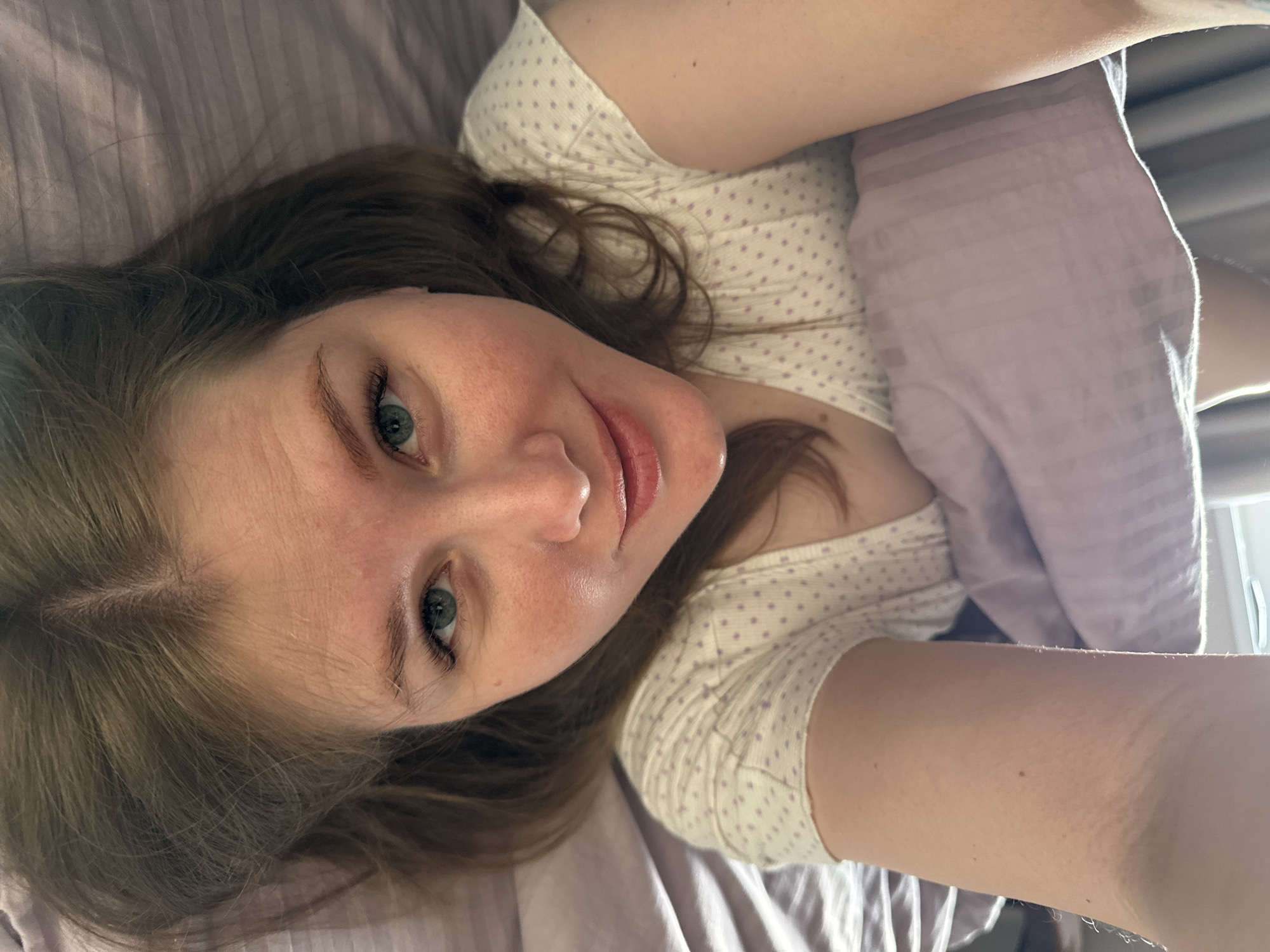 A young woman is captured in a close-up, overhead selfie, lying down in what appears to be a bed. She has long, light brown hair and direct blue-green eyes that engage with the camera. Her lips are slightly parted in a relaxed, gentle smile, conveying a calm and content expression. She is wearing a light-colored top, possibly cream or off-white, adorned with small purple polka dots. A light purple blanket or duvet, featuring a subtle striped texture, partially covers her. A bright light source, likely a window, is visible on the right side of the frame, suggesting it is daytime, possibly morning. The setting appears to be an indoor space, characteristic of a bedroom. There is no visible text in the image. This scene is set in Ферма, Russia.