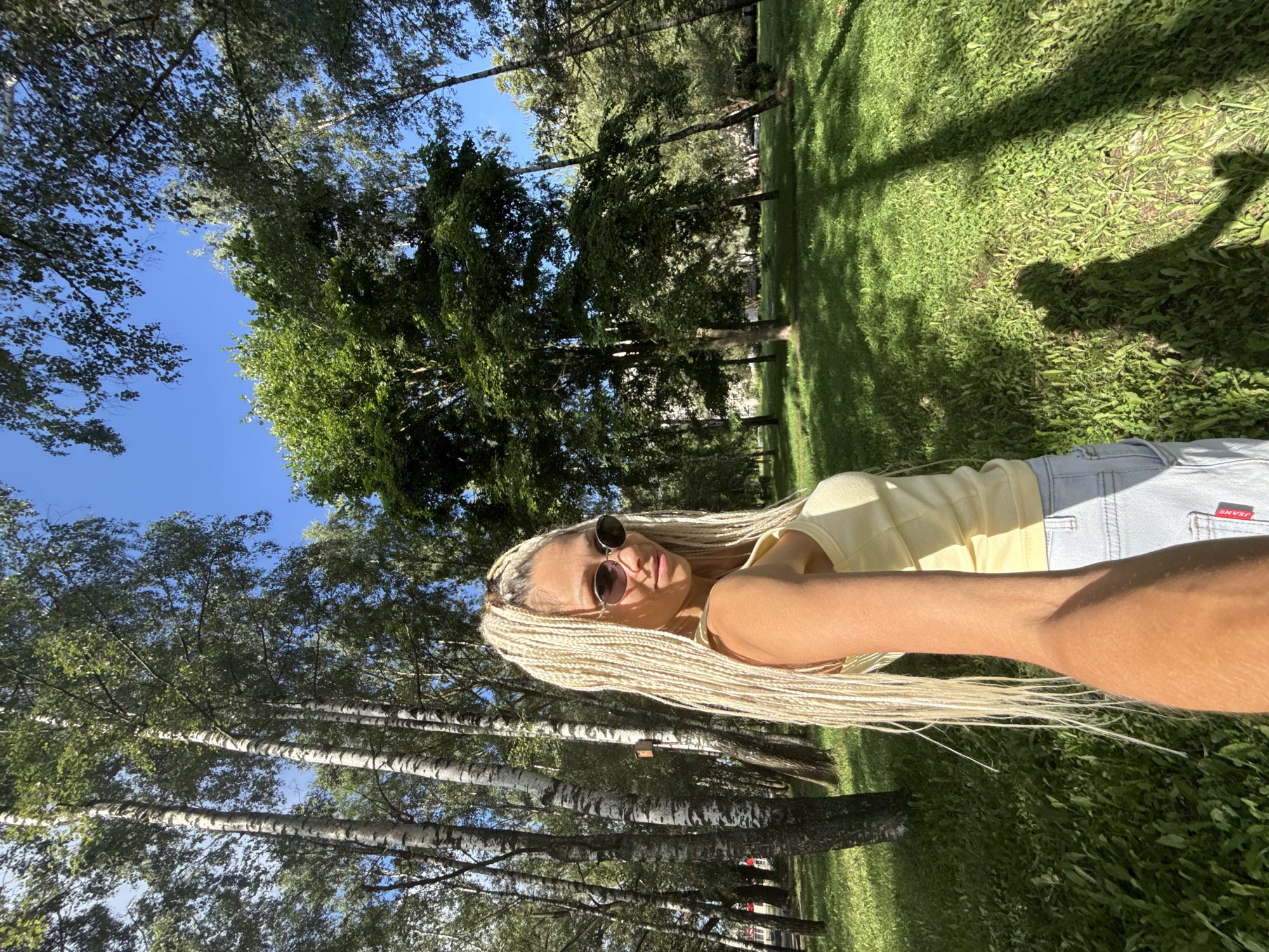 A woman with long blonde braided hair and sunglasses is taking a selfie outdoors in a park with trees and grass under a clear blue sky. She is wearing a yellow tank top and light-colored denim shorts. The woman is positioned in the foreground on the left side of the frame, with her arm extended to take the photo. The background shows a grassy area with trees, and shadows of trees and people are cast on the grass. The overall scene appears to be a sunny day in a natural, urban park setting.