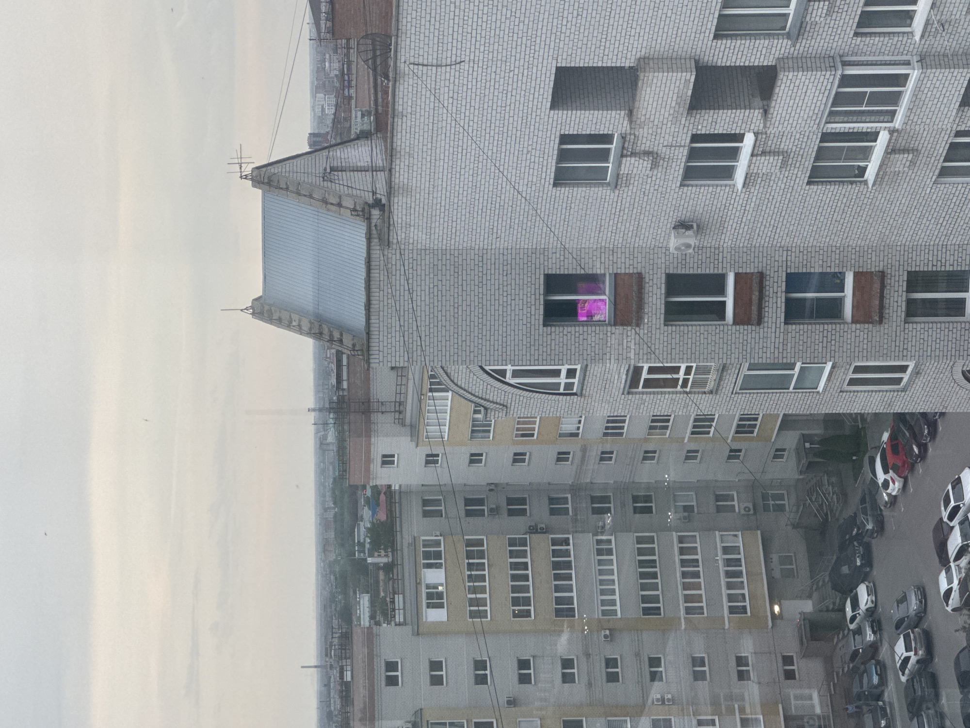 A building in Unknown, Unknown is shown from an elevated perspective. The building has a gray brick exterior with rows of windows. Some windows have open shutters or panels. A smaller building with a metal roof is attached to the side. Below, there are several stories of windows and parking with numerous cars. The weather is overcast.