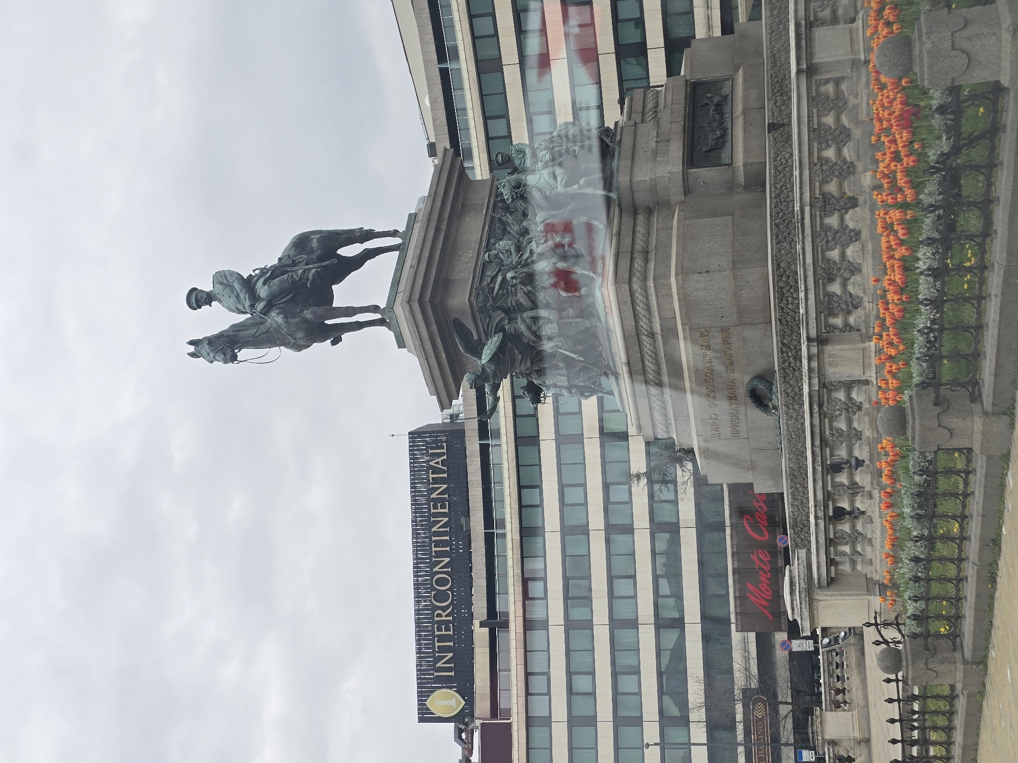 The image captures a view of the Monument to the Tsar Liberator in Sofia, Bulgaria, with the InterContinental Sofia hotel in the background. The monument features an equestrian statue of Emperor Alexander II of Russia and intricate relief sculptures depicting scenes from the Russo-Turkish War of 1877-1878. The base of the monument is adorned with flower beds and surrounded by a wrought iron fence. Street signs, traffic signals and other street furniture are visible near the base. The sky is overcast.