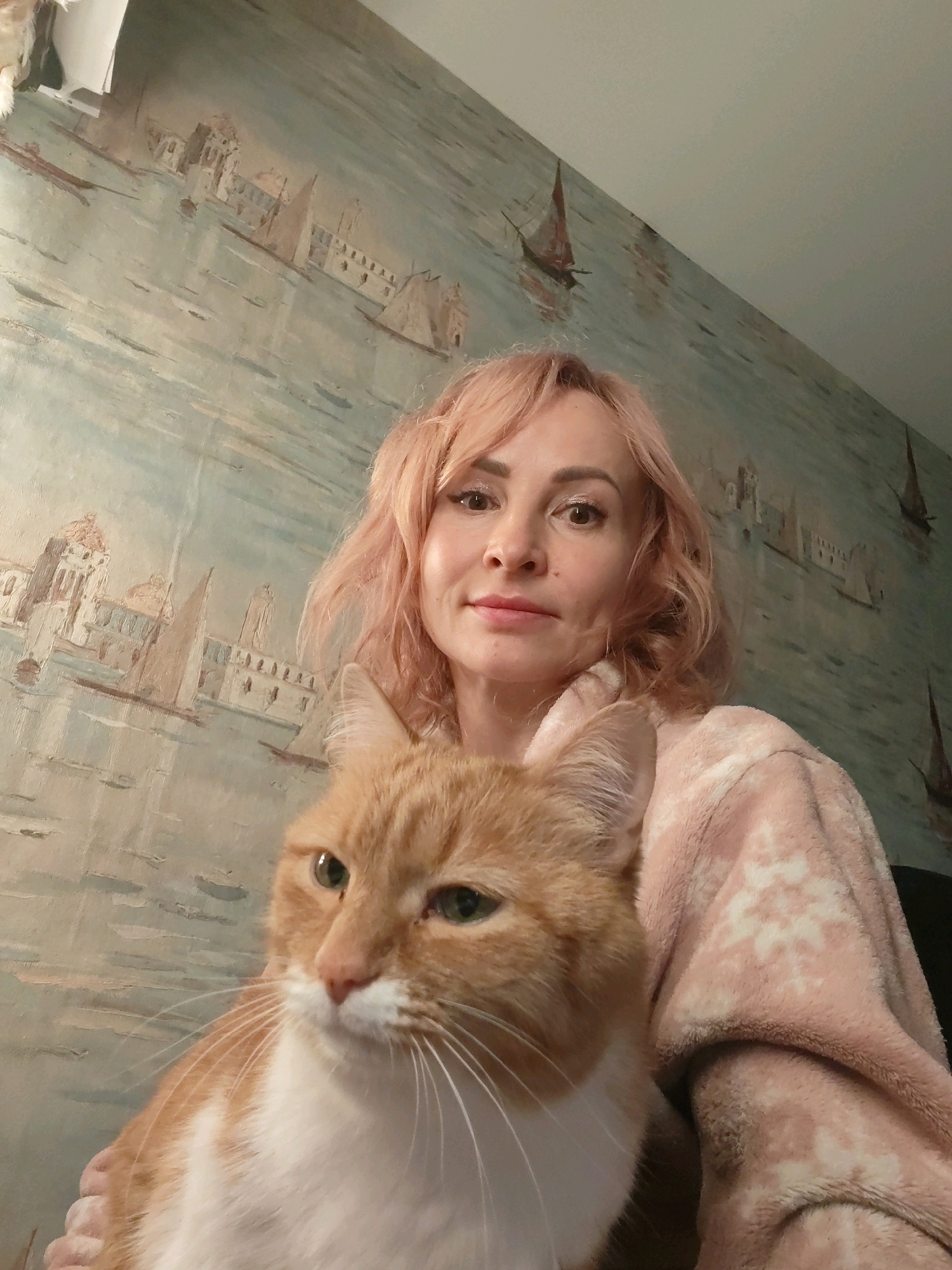 The image is a selfie taken indoors. A woman with short, pink hair and makeup is in the foreground, holding an orange and white cat. The woman is wearing a light pink robe with a snowflake pattern. The cat's face is prominent in the lower left of the frame, with green eyes and white whiskers. Behind the woman and cat is a wall with wallpaper depicting a scenic view of a canal with buildings and sailboats, suggestive of Venice. The overall impression is a relaxed, domestic scene. There is no visual information to suggest the location is the city of Chaikovsky, Russia.