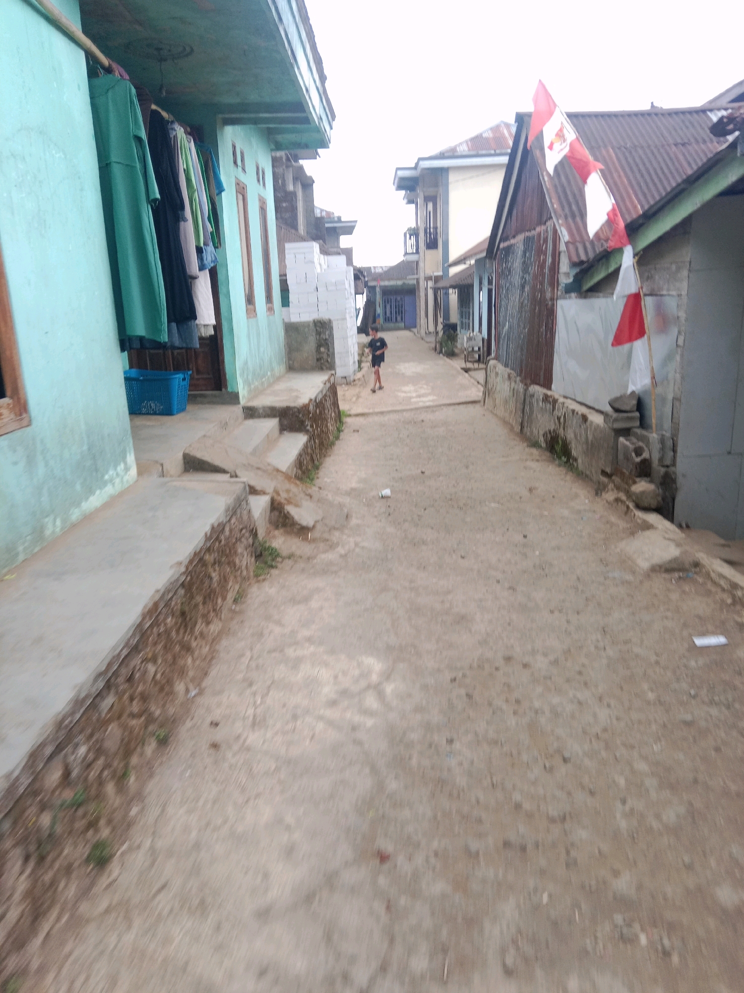 A street scene in Batur, Indonesia, shows a narrow, unpaved path between two rows of buildings. The building on the left is painted a pale turquoise and has laundry hanging from its eaves. On the right, a corrugated metal structure displays a red and white Indonesian flag. A person in dark shorts and a t-shirt is walking away from the camera down the path. The sky is overcast.