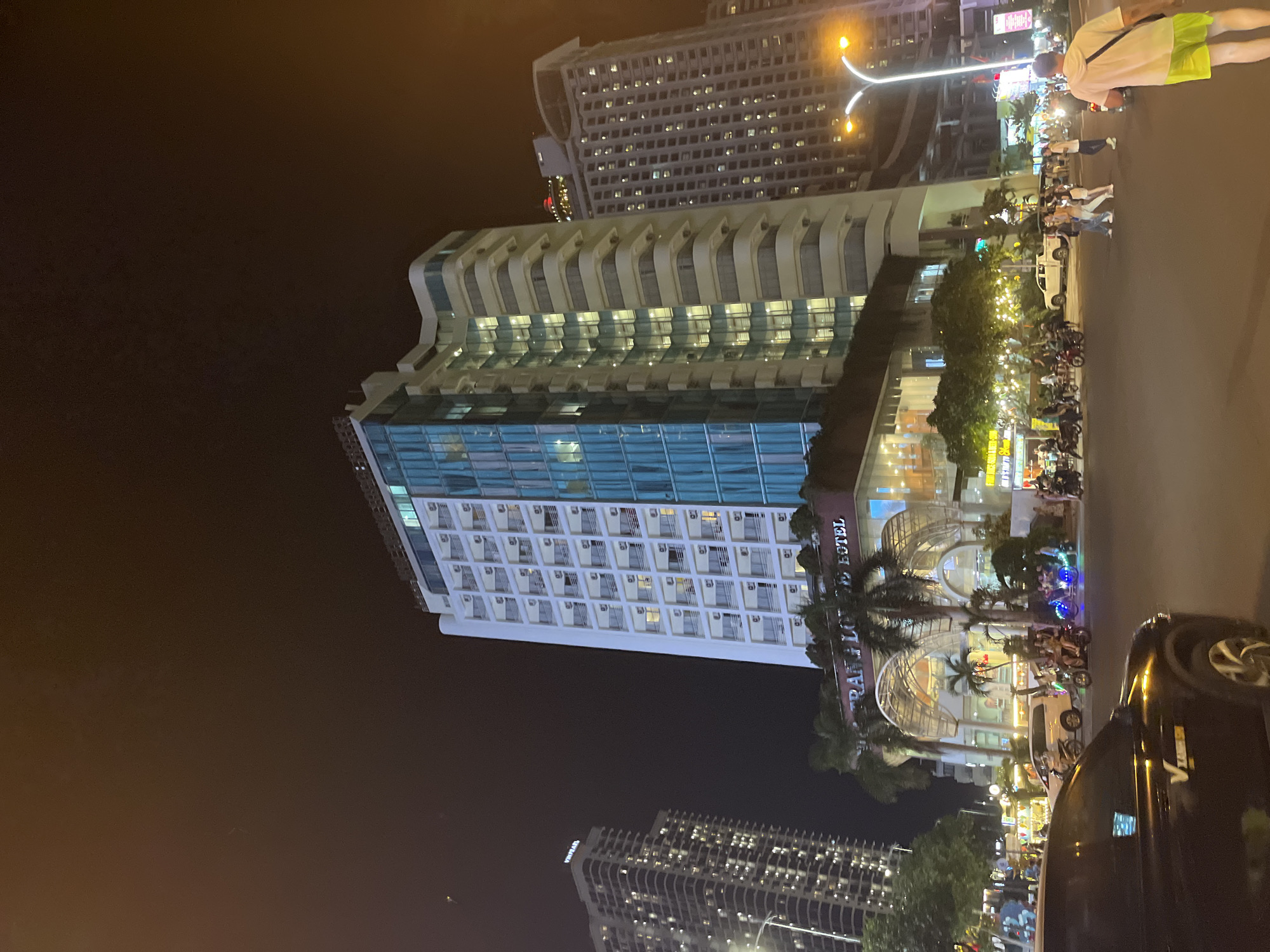 The image depicts a nighttime street scene in Nha Trang, Vietnam, featuring the Tran Phu beach area. In the foreground, a dark-colored vehicle is partially visible. The middle ground showcases a street with various people, motorcycles, and cars. One white van is parked on the right side of the street, and several people are crossing the street. Pedestrians are also seen walking on the sidewalks. The background is dominated by a large, multi-story hotel with balconies and illuminated windows. Adjacent to it, another tall building with many windows is visible. Streetlights illuminate the area, and the sky is dark. The hotel signage reads "TRAN LOI HOTEL." The overall environment appears to be a bustling urban setting.