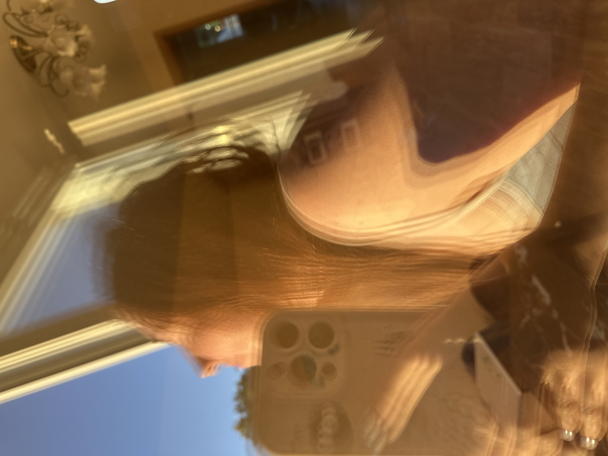 The image is a blurry, motion-filled reflection in a window. A person's head and shoulder are visible, with long, light brown hair. They appear to be looking out of the window. A smartphone is in the foreground, held by the person, and its camera array is visible. In the background, a chandelier with multiple light fixtures is reflected on the ceiling. The scene suggests an indoor setting with a window offering a view of a blue sky and some trees. There is no specific indication of the location being Nizhny Novgorod, Russia.