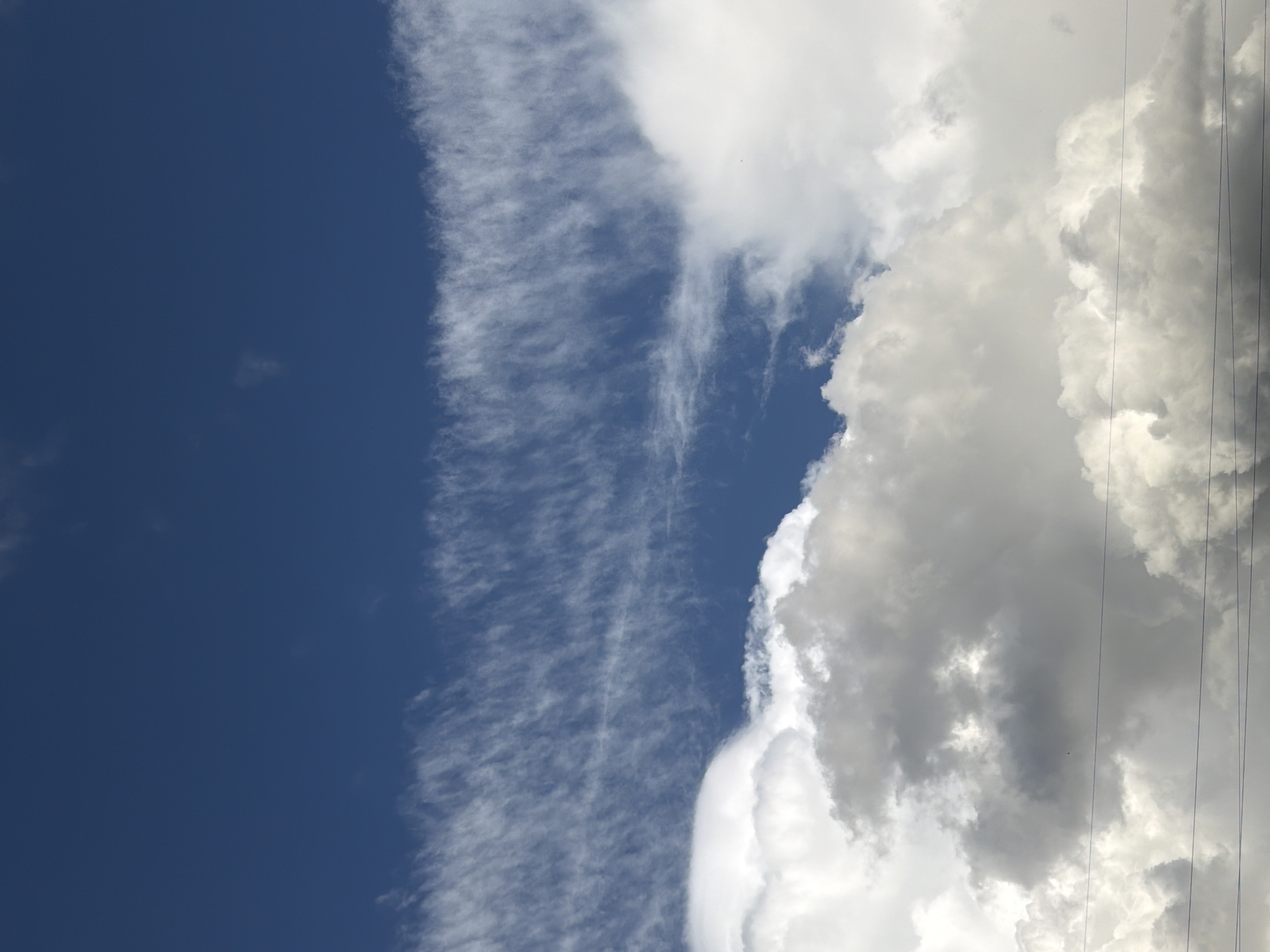 A slightly low-angle shot of clouds in Unknown, Unknown, shows a dark blue sky on the left, a narrow band of wispy clouds to the right of the blue, and then a dense mass of white and gray clouds on the right. The scene is lit by bright sunlight. Some thin electrical wires are visible in the extreme upper-right corner.