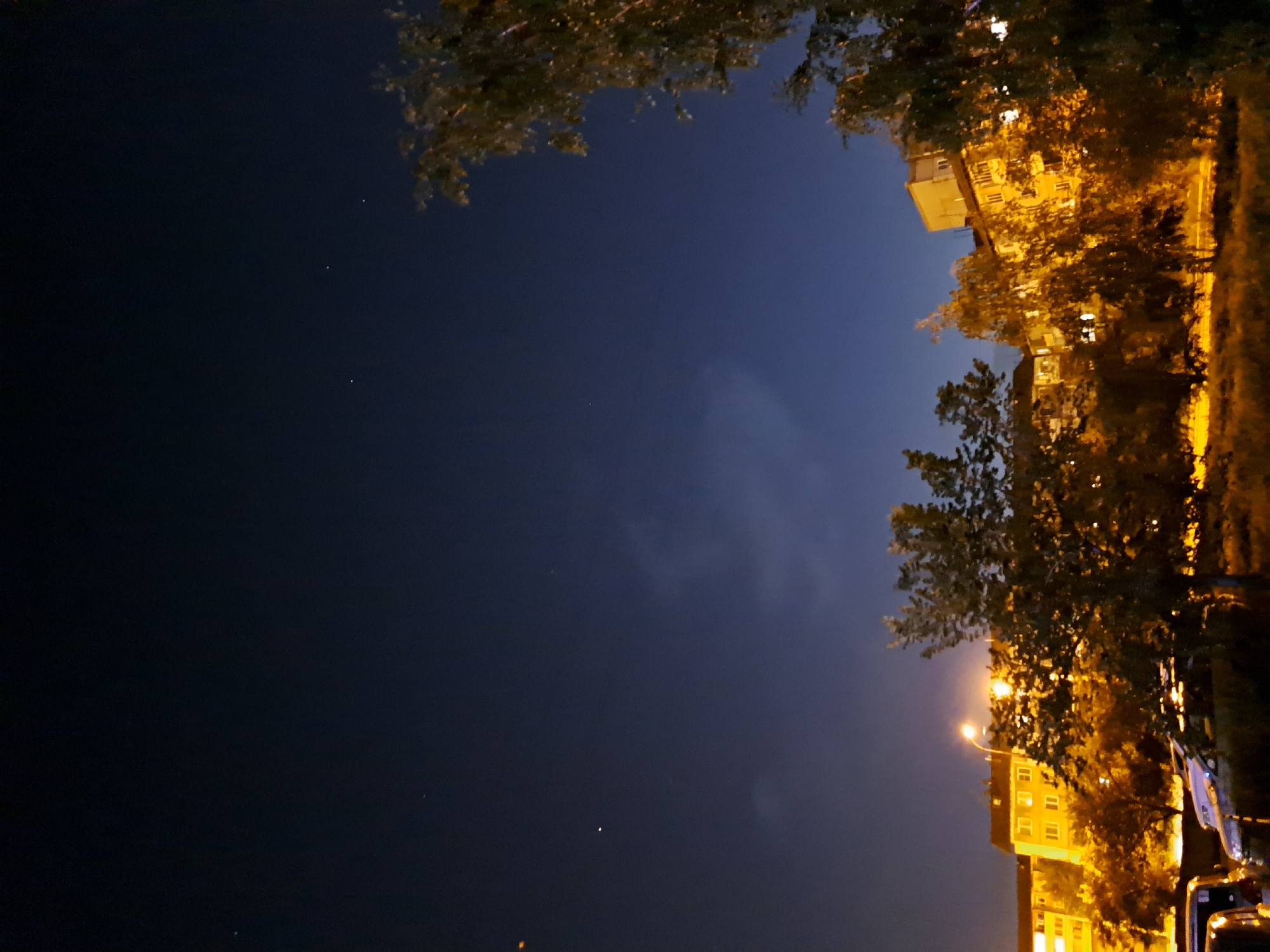 The image depicts a nighttime scene in a residential area, likely in Irkutsk, Russia, with apartment buildings illuminated by streetlights and interior lights. Trees with foliage are scattered throughout the foreground and midground. The sky is dark blue, with a few faint stars visible. In the foreground, parts of vehicles are visible. The overall impression is one of a typical urban evening.