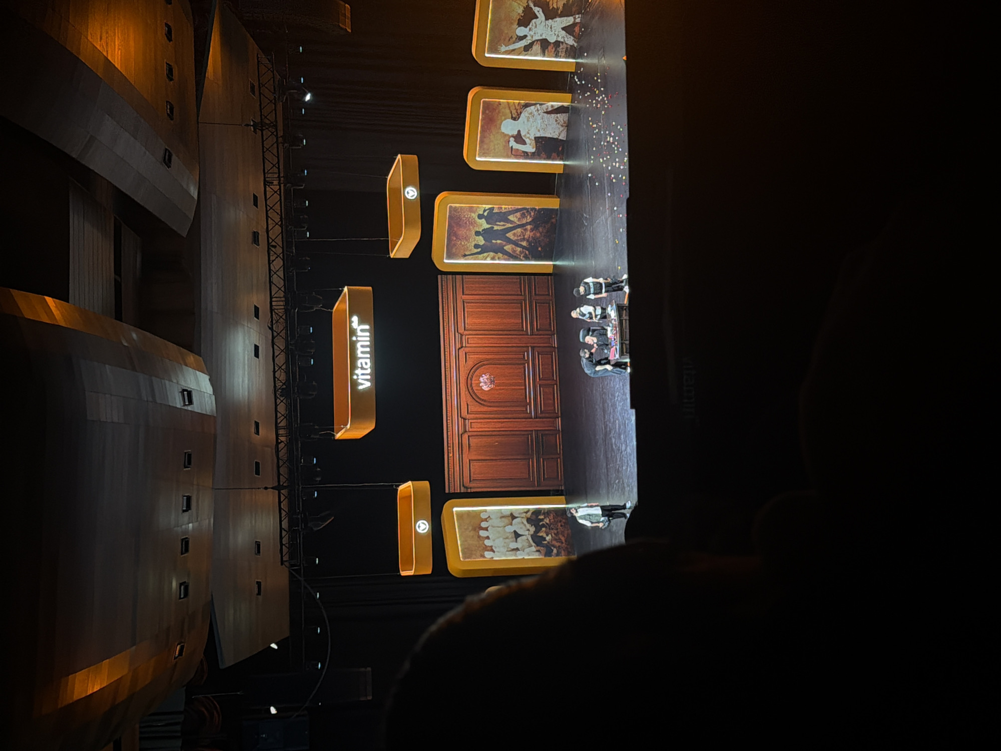 The image depicts a stage performance, possibly at a theater or concert hall in Yerevan, Armenia. The stage is set with an ornate wooden backdrop resembling large doors. Illuminated rectangular displays, some bearing the word "vitamin," are positioned on either side of the stage. These displays exhibit images of figures and scenes. Several people, presumably performers, are gathered near the center of the stage. The lighting is focused on the stage, creating a stark contrast with the surrounding darkness. The image is taken from an audience perspective, with the back of someone's head visible in the foreground.