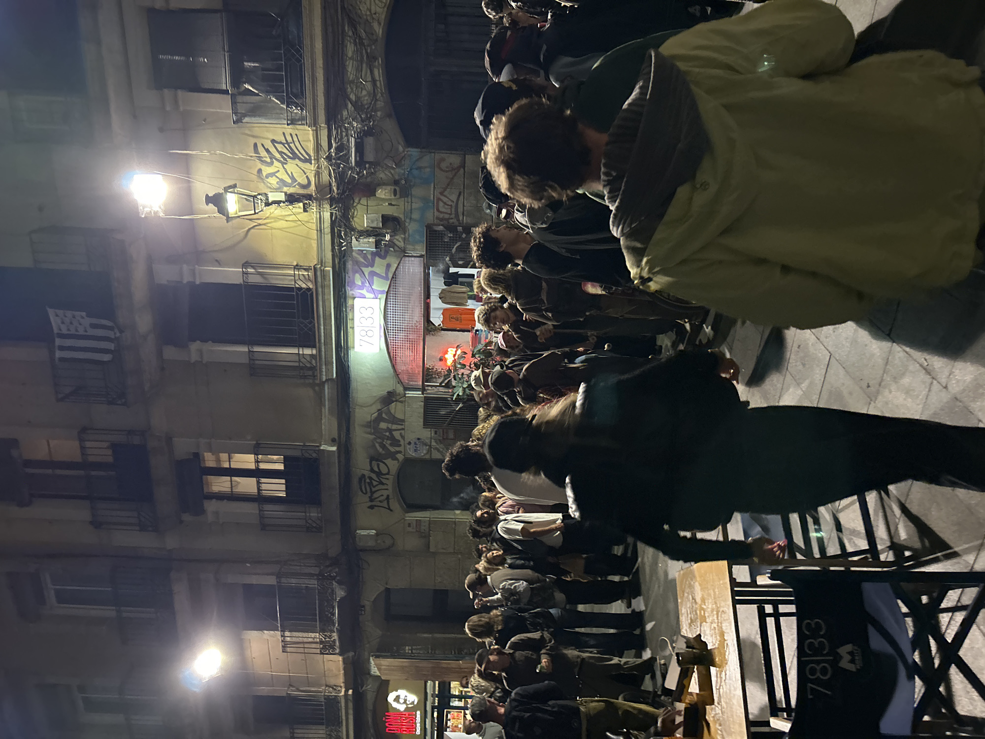 This image captures a vibrant, crowded street scene at night in an urban environment, identified as Barcelona, Spain. Numerous people are gathered along a narrow street or alley, mostly standing with their backs or sides to the viewer, suggesting they are observing an event, socializing, or queuing.
On the left side, multi-story buildings with characteristic European architecture, including windows, iron grilles, and balconies, line the street. Graffiti is visible on the building walls. Bright streetlights illuminate the scene, casting strong light and shadows, and a red glow emanates from a shop or sign further down the street. A black and white flag with horizontal stripes and a canton, resembling the Breton flag (Gwenn ha Du), hangs from one of the buildings.
In the foreground on the right, parts of a wooden table and black chairs are visible. The crowd appears dense, indicating a lively and social atmosphere.
Visible text includes the numbers "78|33" on a prominent sign above a shop entrance, which also appears on a black fabric item (possibly a chair back or bag) in the bottom right corner, accompanied by a small "M" logo. Another shop sign partially visible reads "Dona Rush". The time of day is clearly night, as evidenced by the artificial lighting and overall darkness.