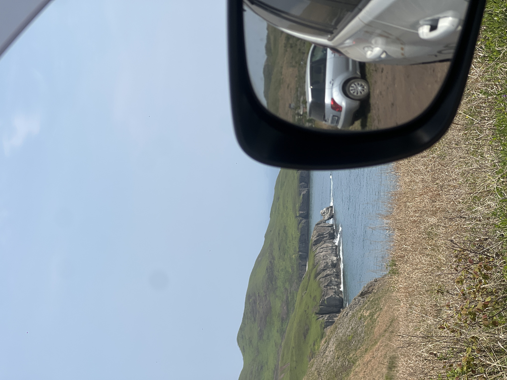 The image depicts a coastal scene with a body of water bordering a green, hilly landscape. A vehicle side mirror is prominent in the foreground, reflecting another vehicle parked on a dirt surface. The location is identified as Slavyanka, Russia. The environment includes a clear sky and vegetation that appears to be grassy or shrub-like near the viewer's location. The coastal area shows rocky cliffs meeting the water's edge.