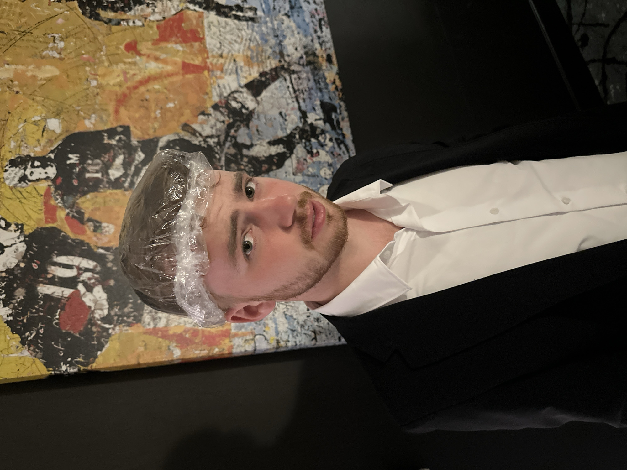 A person with light skin, short brown hair covered by a clear plastic shower cap, a neatly trimmed beard, and green eyes is wearing a white dress shirt with visible buttons and a black suit jacket. The person is positioned in front of a textured wall or artwork that features a mix of yellow, orange, black, and white colors. The background is dark, and the scene is lit by an unseen light source. The specific location within College Park, United States of America cannot be determined from the image alone.
