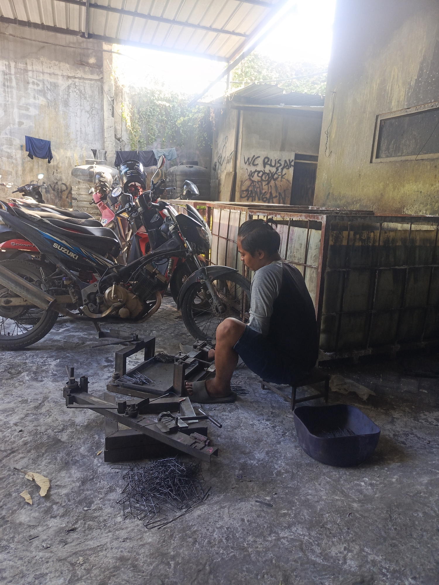 In Sidowungu, Indonesia, a young male, with dark hair pulled into a bun, sits on a low stool in a cluttered open-air workshop. He wears a grey long-sleeved shirt and dark shorts, with one foot clad in a sandal visible. The workshop environment is characterized by a concrete floor, a partially covered roof with corrugated metal, and rough concrete walls, one of which is covered in graffiti. Several motorcycles are parked haphazardly in the background. In the foreground, a collection of metal objects, possibly fasteners or wire, is scattered on the floor and arranged on a metal fixture. A dark, oval-shaped container sits nearby. The scene is illuminated by bright sunlight filtering through the roof.