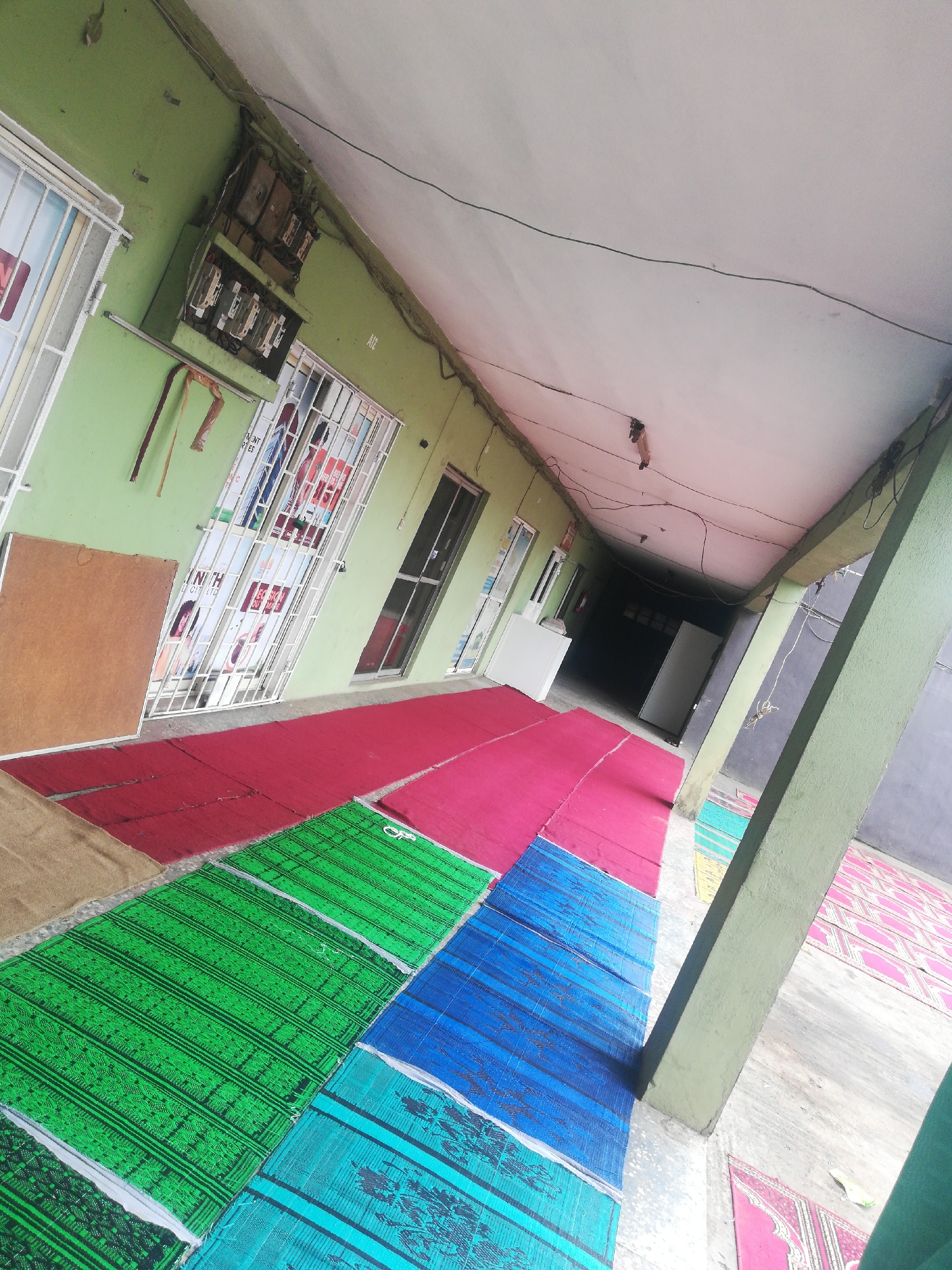 The image shows a covered outdoor corridor in a building, possibly in Unknown, Unknown. The building has light green walls and a white ceiling. Electrical boxes and wiring are visible on the wall. A series of colorful mats, including green, blue, and red, are laid out on the floor of the corridor. Windows or openings with bars are present on one side of the corridor, displaying posters or advertisements. A few doors are also visible along the corridor. The far end of the corridor leads into a darker interior space. A support column is seen on the right side.