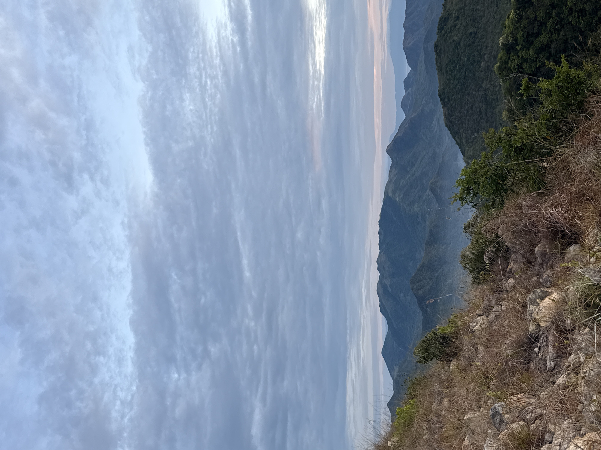 The image shows a landscape in what appears to be the Bac Nha Trang Ward area of Vietnam. The foreground features dry, rocky terrain with sparse vegetation and some green shrubbery. In the midground, a series of mountains fade into the distance, with the closest ones covered in dense green foliage. The background is dominated by a cloudy sky with a hint of sunrise/sunset hues. The image is taken from an elevated vantage point.