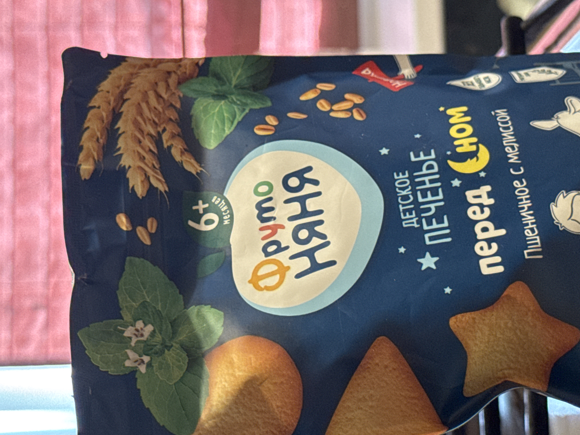 This is a close-up, angled shot of a blue package of baby cookies, likely taken indoors. The packaging features various cartoon illustrations of animals, a crescent moon, and stars, suggesting a nighttime or bedtime theme. Prominently displayed on the package is the brand name "ФрутоНяня" in large, stylized Cyrillic letters. Below this, in smaller Cyrillic text, it reads "ДЕТСКОЕ ПЕЧЕНЬЕ" (Children's Cookies) and "с мелиссой" (with melissa). There is also a marking "+6 месяцев" (+6 months), indicating the recommended age for consumption. Scattered across the blue background are small illustrations of oats or grains, sprigs of mint with small white flowers, and various shapes of cookies, including star-shaped ones. The overall impression is of a product aimed at young children, emphasizing natural ingredients and a gentle, comforting theme. The background shows a blurred pink and white striped curtain, suggesting an indoor setting, possibly a home.