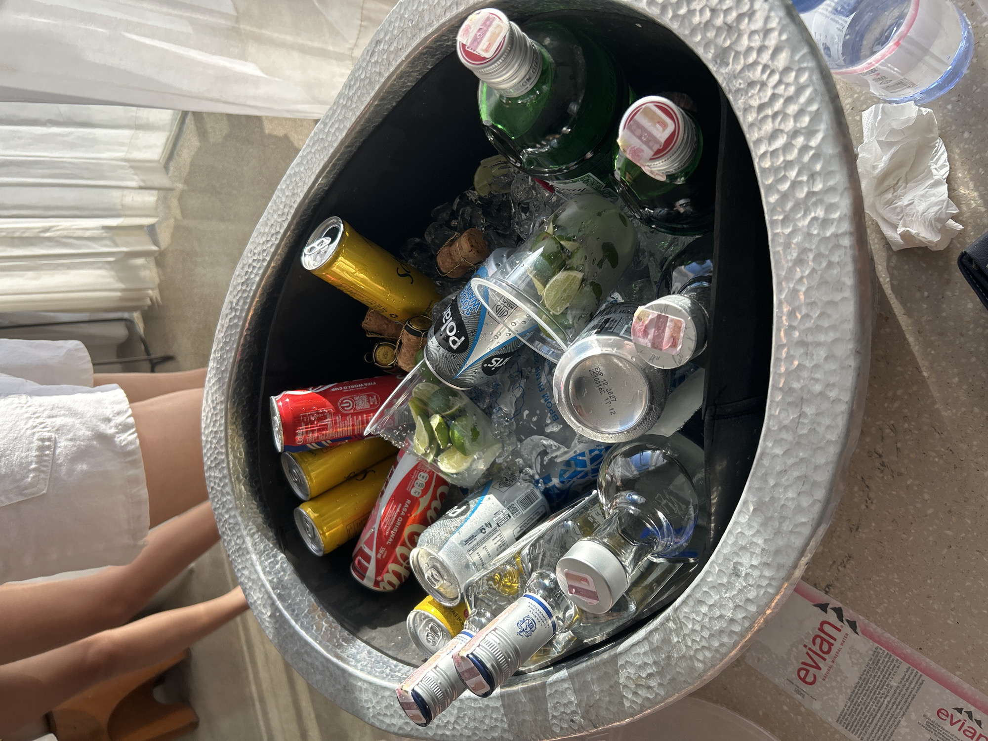 The image shows an outdoor scene in North Kuta, Indonesia, with a hammered metal ice bucket filled with various beverages and ice. The bucket contains several cans of soda, including Coca-Cola and what appear to be other carbonated drinks, along with bottles of clear spirits, a green bottle of liquor, and several plastic cups with lime slices. Some corks from wine or champagne bottles are also visible. In the background, part of a person's legs wearing white shorts is visible, and sheer white curtains hang in front of a window. The ground appears to be a speckled terrazzo or concrete surface, with a discarded Evian water bottle and a crumpled tissue nearby. The lighting suggests it is daytime, possibly late afternoon, with sunlight casting shadows.