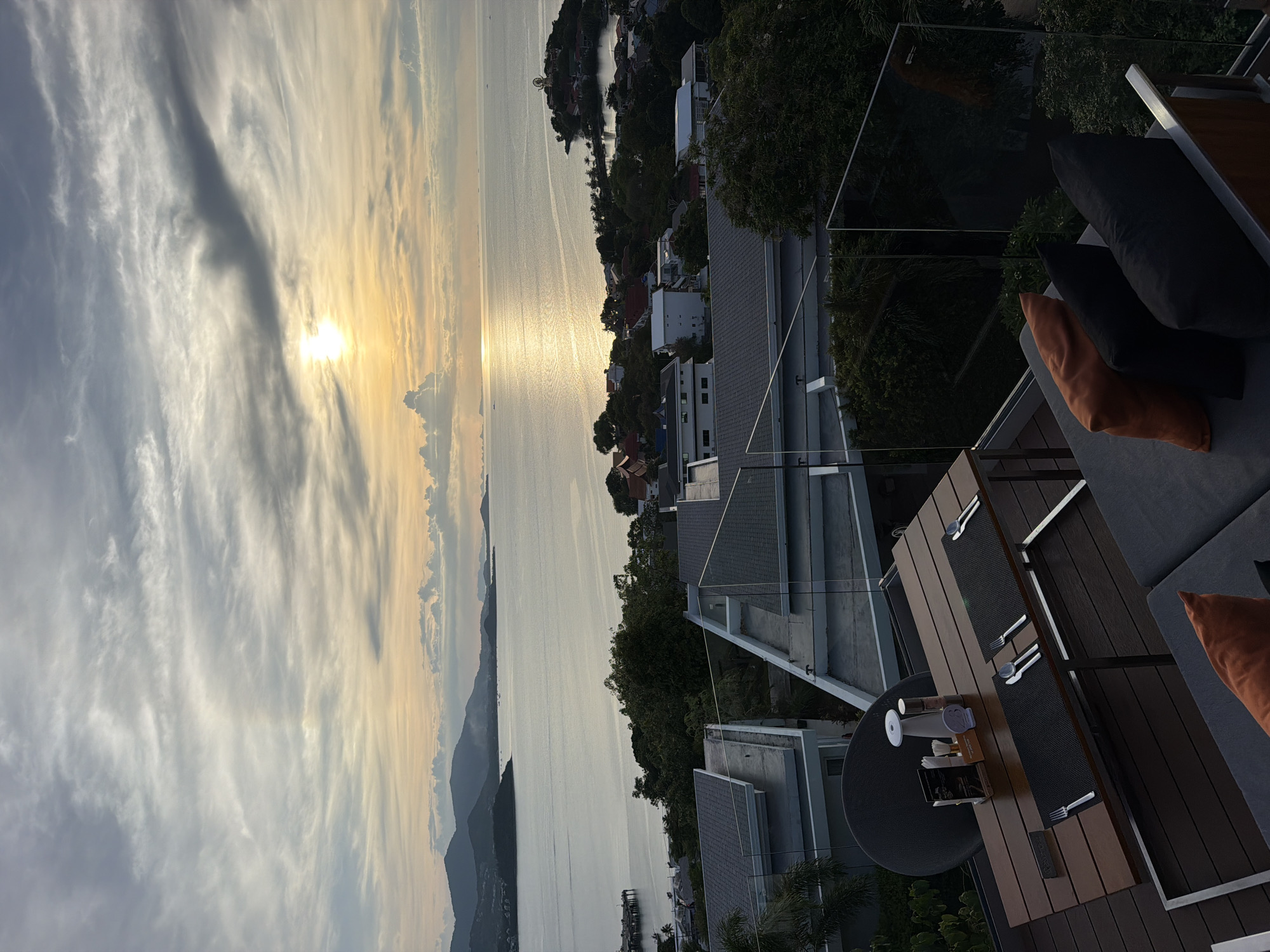 This is an eye-level, long shot, vertical photograph of a rooftop dining area overlooking a tropical seascape at dusk. The sun is low in the sky, casting a golden sheen across the calm water and illuminating a scattering of clouds. In the foreground, a wooden table is set for dining with placemats, cutlery, and decorative items. A lounge sofa with several cushions is positioned nearby. Beyond the immediate dining area, a cluster of modern buildings with grey roofs is visible, interspersed with lush green trees. Further in the distance, rolling hills and the vast expanse of the ocean stretch towards the horizon. The scene is located in Baan Plai Laem, Thailand.
