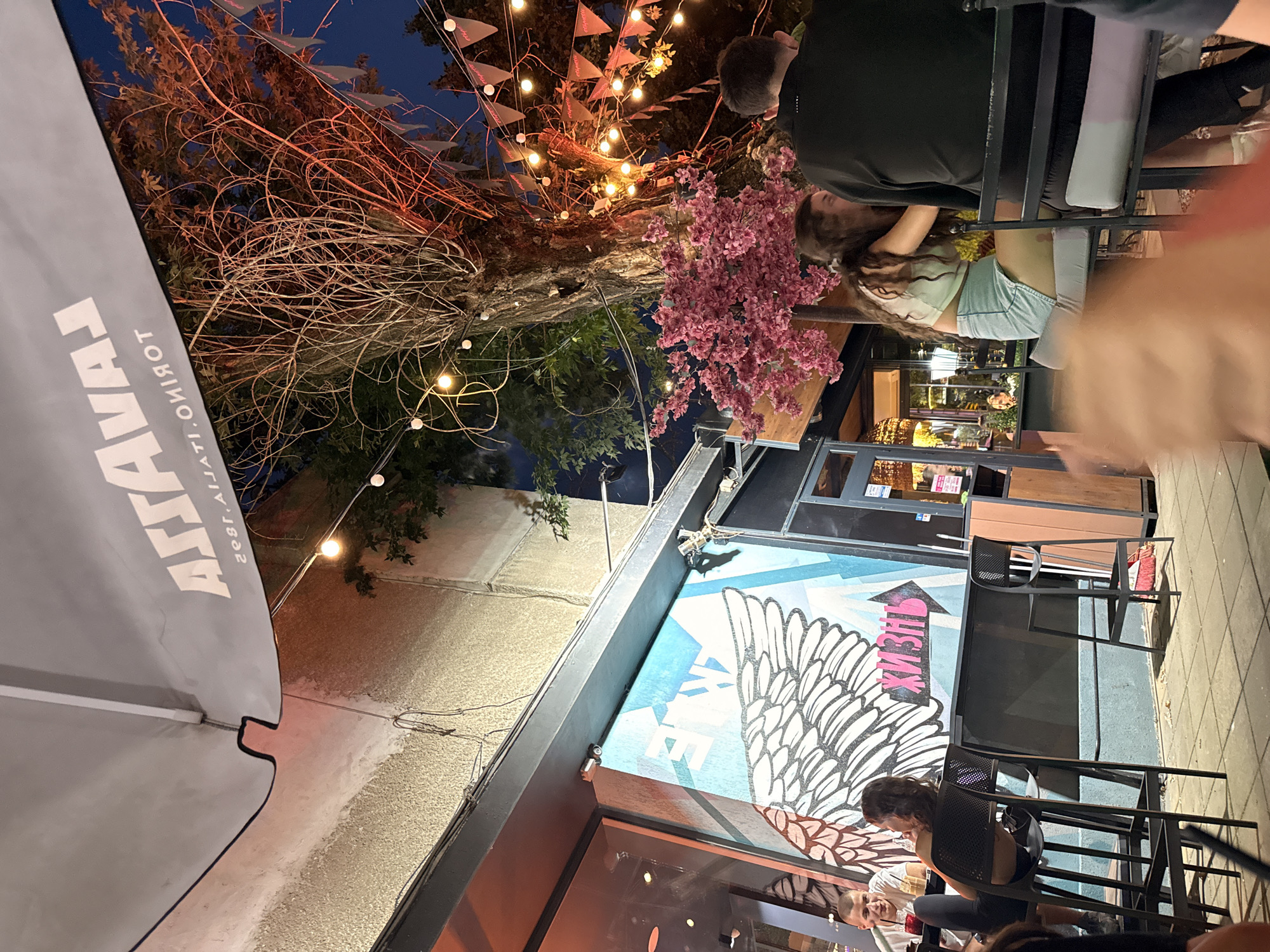 The image depicts an outdoor seating area at night, possibly a cafe or restaurant, located in Lovech, Bulgaria. A large grey umbrella with the "LAVAZZA" logo is visible at the top. String lights and decorative flags are strung across a tree and the surrounding area, illuminating the scene. In the foreground, two individuals are seated at a table, facing away from the camera. A mural featuring large white wings and the word "Жизнь" (Life) in pink lettering is painted on the wall behind them. The overall atmosphere is dimly lit and appears to be a casual outdoor dining or social setting.