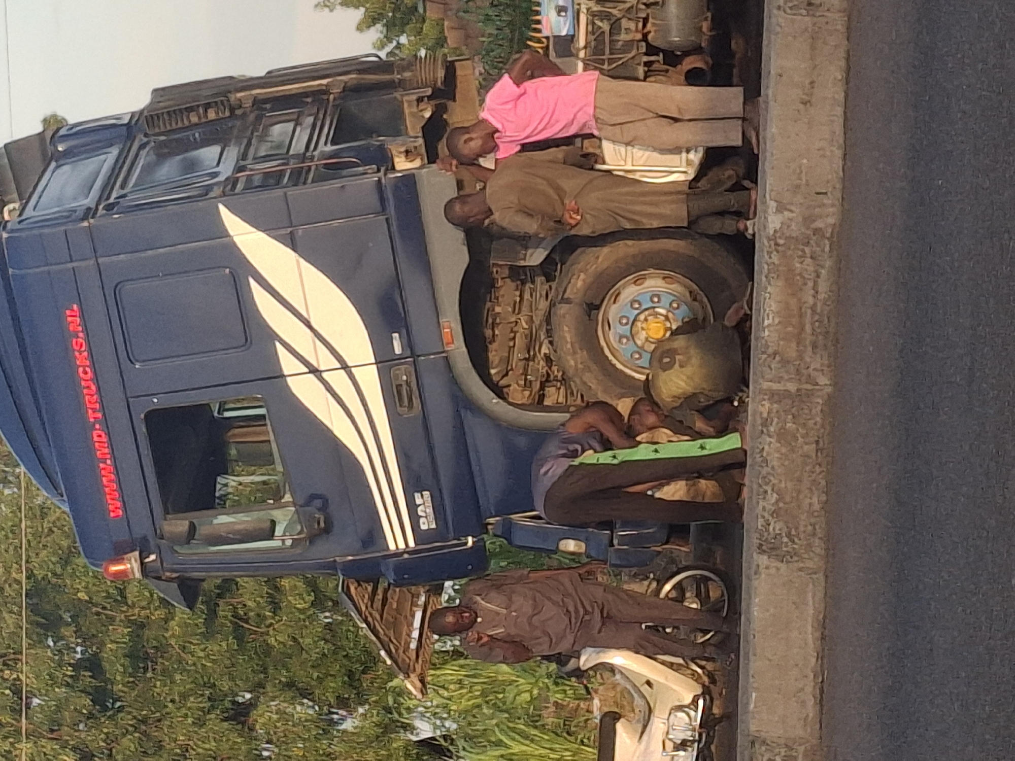 A semi-trailer truck is stopped on the side of a road in Gombe, Nigeria, with its front tilted upwards at an angle. Four individuals are gathered around the front of the truck, appearing to be inspecting or working on it. One person, wearing a grey shirt and pants, stands and gestures with their hand. Another person, in a blue shirt and dark pants, is crouched down. Two other individuals, one in a brown robe-like garment and another in a pink shirt and beige pants, are standing near the truck. A white motorcycle is parked to the left of the truck, and lush green foliage is visible in the background. The road surface appears to be asphalt, with a concrete curb separating it from the area where the truck is stopped. The sky is visible at the top of the frame, suggesting an outdoor setting, likely during daylight.
