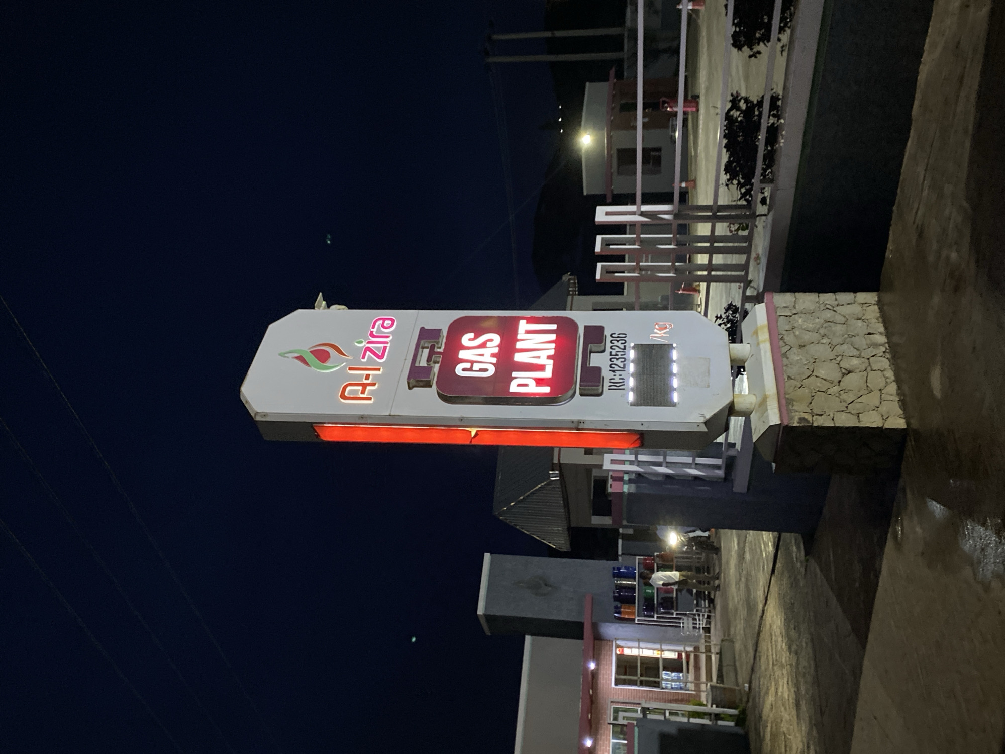 The image depicts a gas plant sign at night in Jos, Nigeria. The sign is illuminated and features the text "Al-Zira GAS PLANT" prominently. Below this, there is a reference number "RC-1235236" and a measurement indicating "/Kg". The sign is white with colorful elements and red and pink accents. In the background, there are buildings, some with lighting, and structures that appear to be part of the gas plant or surrounding facilities. The sky is dark, suggesting it is nighttime. The ground is wet, indicating recent rain or damp conditions. The overall scene suggests a commercial establishment operating in a town or city at night. There are no visible people in the immediate vicinity of the sign, but the background suggests some activity in the buildings. The lighting from the sign and the buildings creates a somewhat atmospheric night scene.