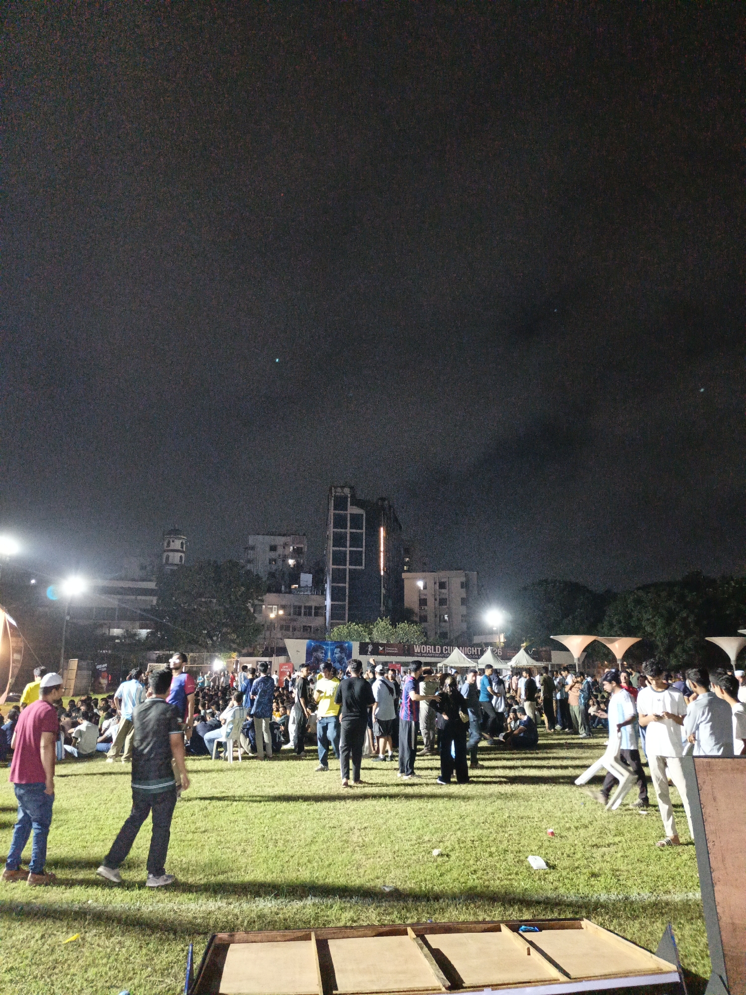 The image shows an outdoor nighttime gathering of a crowd of people on a grassy field. In the background, there are several buildings, including a multi-story modern building with a grid pattern of windows and a more traditional building with a domed tower. Several bright stadium lights illuminate the scene from the left side. A large banner with text that reads "WORLD CUP NIGHT" is visible in the middle background. Many people are standing and milling about, while some are seated. The ground is covered in green grass, and there are some wooden structures in the foreground, one of which appears to be a low platform with dividers. The sky is dark and cloudy, suggesting it is nighttime. This event is taking place in Dhaka, Bangladesh.