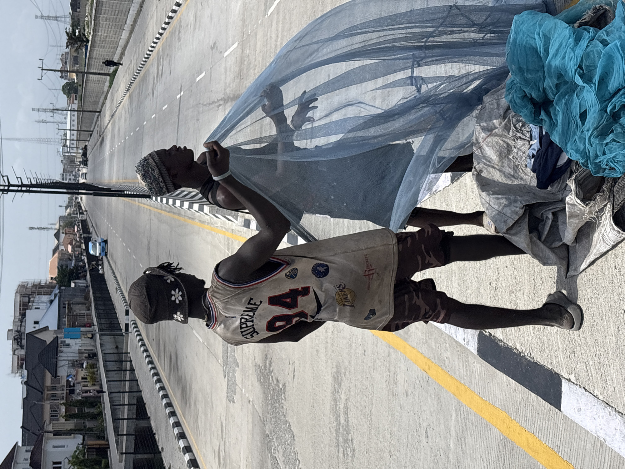 Two individuals are depicted standing on the side of a wide, modern road or bridge under bright daylight in Igboefon, Nigeria. The image is oriented vertically (rotated 90 degrees clockwise).
One individual, positioned closer to the viewer, wears a light-colored, possibly crocheted beanie and a dark sleeveless top. They are holding up a large piece of translucent blue mesh fabric, which billows and stretches across the frame, partially obscuring the second person. Their hands are visible grasping the fabric.
Behind them stands another individual, wearing a beige sleeveless jersey adorned with various logos, including "Supreme 84," "Lakers," "Houston Rockets," and a Nike swoosh. They also wear camouflage-patterned shorts and a dark cap decorated with white flower patterns. Bundles of other fabrics, including a crumpled blue mesh and gray textiles, are piled on the ground beside them.
The road features clear white dashed lines, solid yellow lines, and black-and-white striped barriers along its edges. In the background, a sprawling cityscape unfolds with numerous residential and commercial buildings. Tall power lines and transmission towers stretch across the horizon. A light blue car is visible in the distance on the road. The clear sky and strong shadows suggest a sunny day. The scene captures an everyday moment along a busy-looking infrastructure.