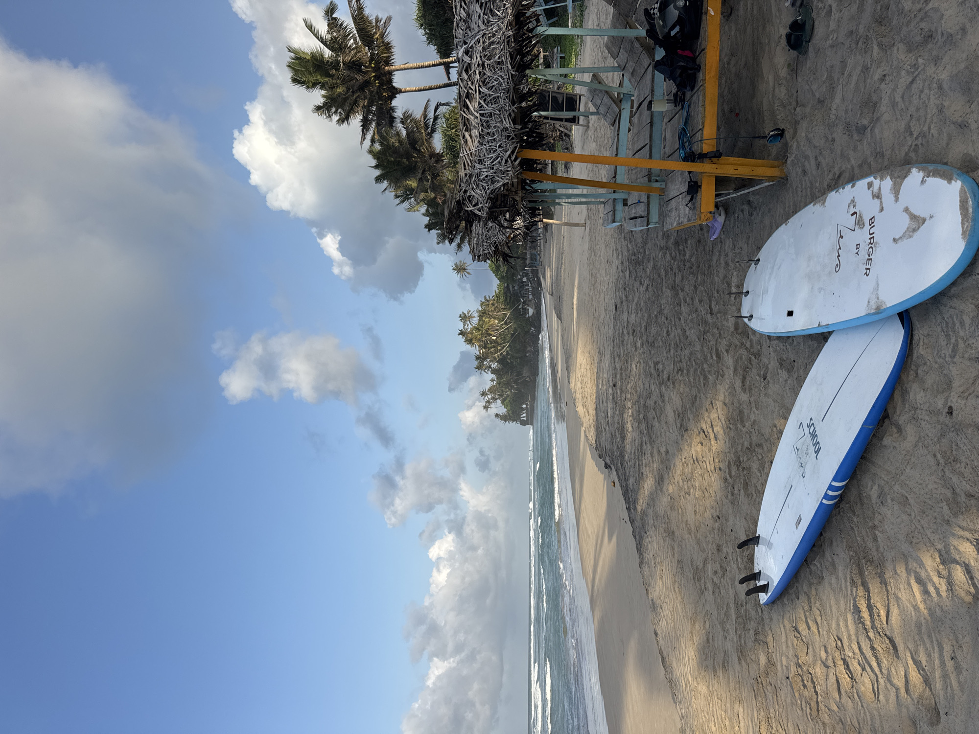This is a photograph taken at a beach in Kathaluwa, Sri Lanka. The image captures a serene beach scene with two surfboards lying on the sand in the foreground. The surfboards are white with blue accents and fins. In the middle ground, there are several thatched-roof beach cabanas with wooden lounge chairs underneath. Palm trees are visible throughout the scene, with some in the middle ground and more densely packed in the background on a slightly elevated area. The ocean stretches across the background, with waves gently rolling towards the shore. The sky is a mixture of blue with scattered white and gray clouds, suggesting it might be early morning or late afternoon. The sand is light brown and appears to have been recently raked or patterned by the wind. There are no people visible in the image.