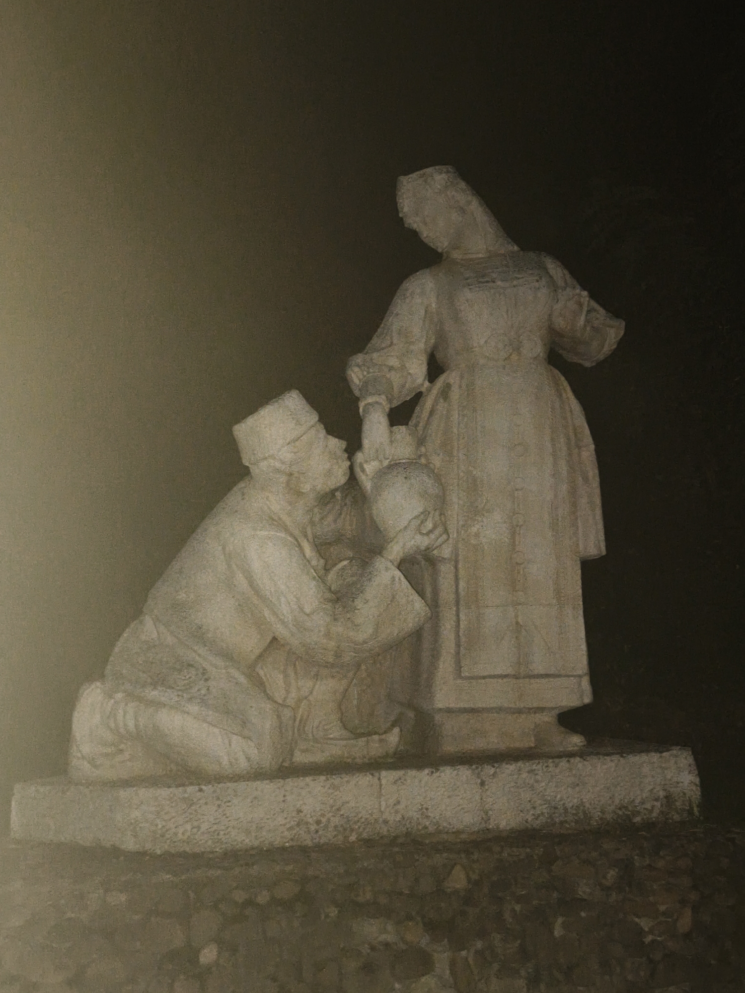 The image shows a stone sculpture of two figures in an outdoor setting. The sculpture is of light-colored stone and appears to be weathered. On the left, a male figure is kneeling, wearing a hat and traditional clothing. He is looking up at a female figure who is standing. The female figure is also wearing traditional clothing and has a long dress. She is holding a large jug from which she appears to be pouring liquid into a container held by the kneeling male figure. The sculpture is placed on a raised stone platform, which is in turn situated on a base of rough stones. The background is dark and indistinct, suggesting a night scene or a poorly lit area. The overall impression is of a scene depicting a communal activity or a moment of service. This sculpture is located in Lovech, Bulgaria, specifically in the central area of the town, likely in a park or public square.