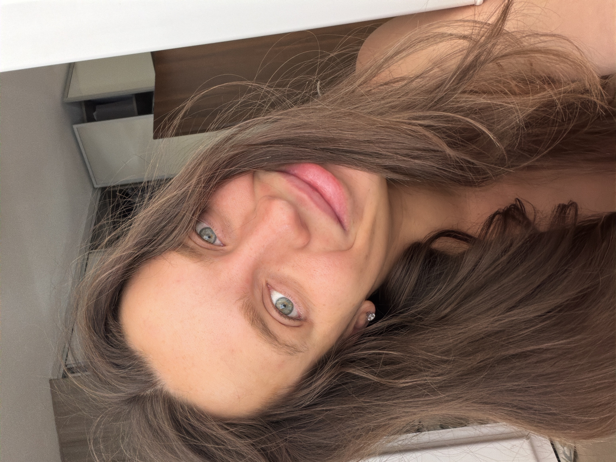 A close-up selfie of a woman with long, brown hair, light skin, and light blue eyes is shown. She is smiling slightly. She appears to be indoors, with a white shelf or structure visible in the upper portion of the image. The environment also shows what appears to be white drawers. No specific information indicates this image was taken in Budva, Montenegro. No interactions or specific actions are observable in the photograph, beyond posing for a selfie.