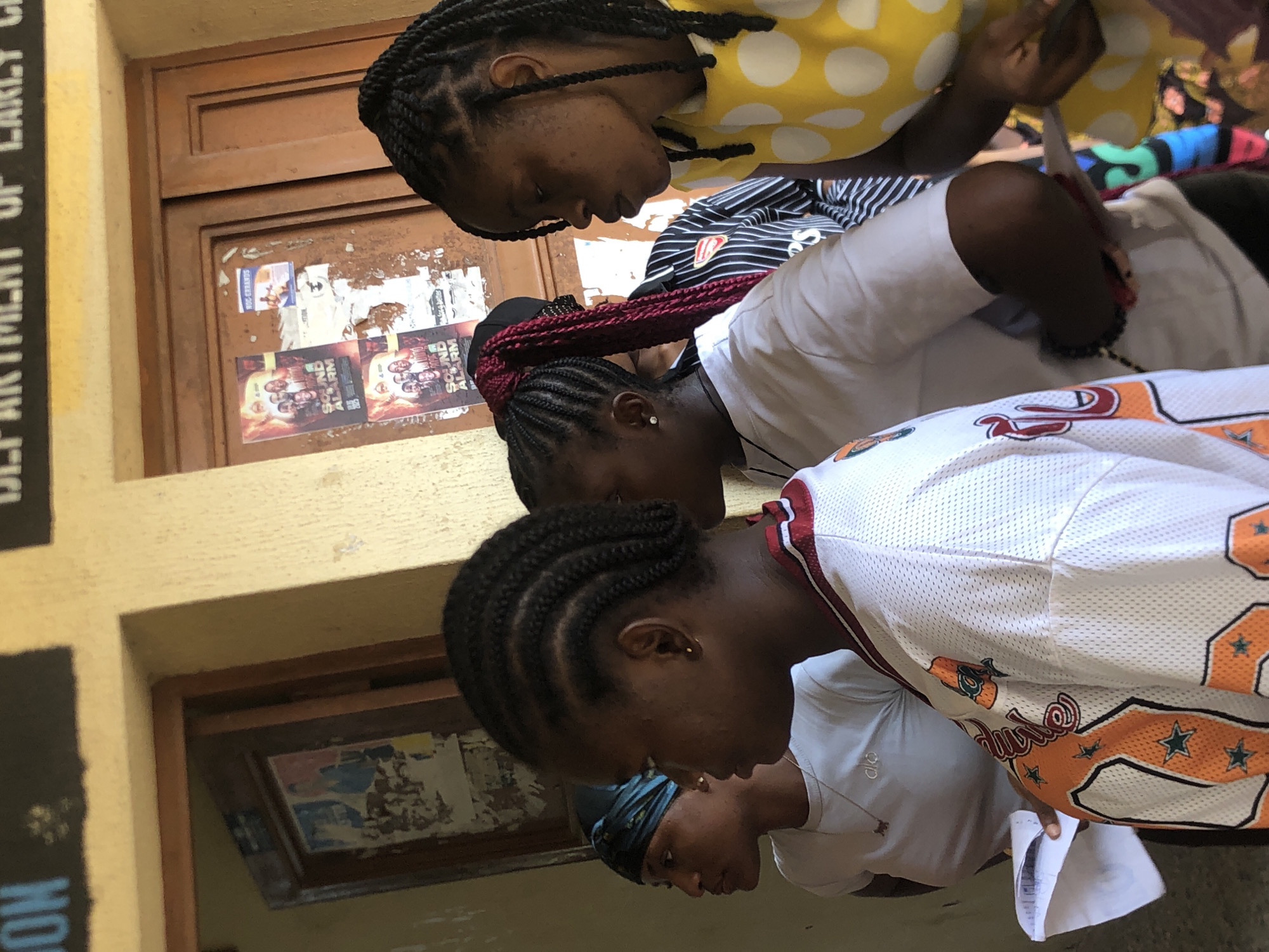 In Ukwu-Abwa, Nigeria, an outdoor shot shows a group of at least four young women focused on something together. One wears a yellow polka dot shirt, another a white shirt with black stripes and an Arsenal logo. The woman in the foreground is wearing a white basketball jersey. Their attention seems to be on something the first woman is holding. They are standing near a building with posters on the wall. A sign reading "DEPARTMENT OF EARLY CHILDHOOD EDUCATION" is partially visible.