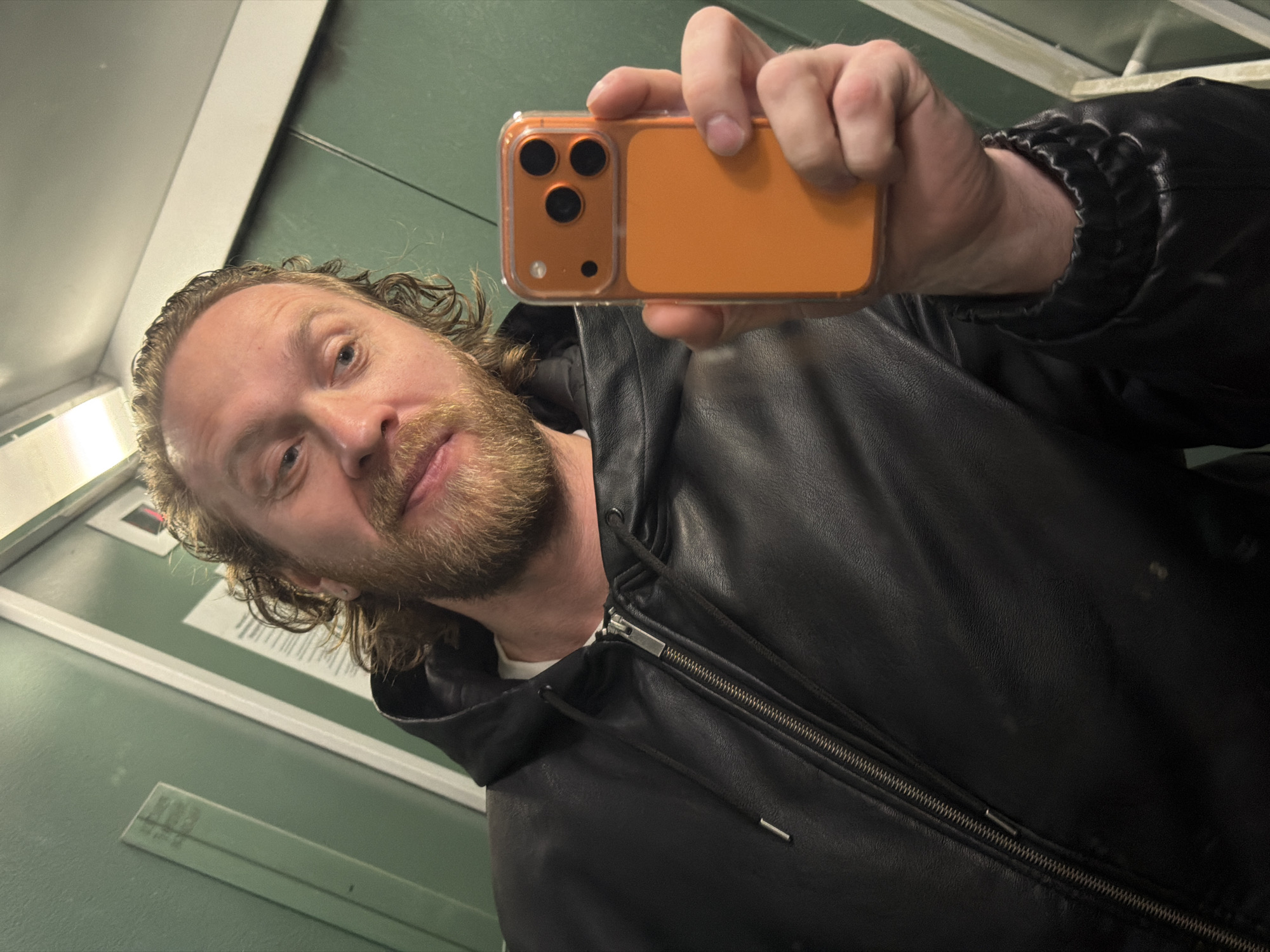 A man with blond hair and a beard takes a selfie with an orange iPhone in what appears to be an elevator with green walls. He's wearing a black leather jacket with a zipper. The location is not discernible from the image.