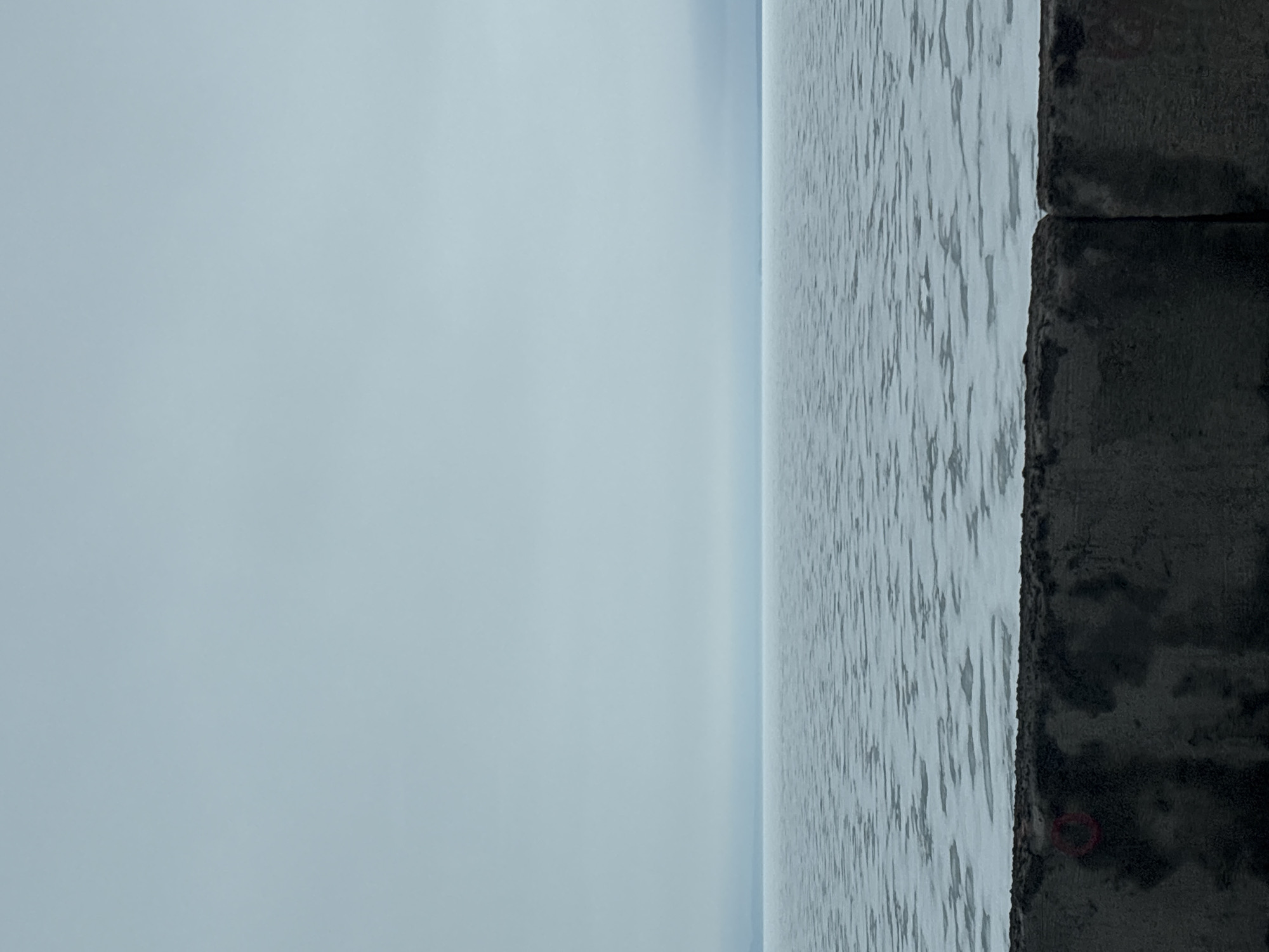 The image shows a body of water meeting a concrete embankment in Chaikovsky, Russia. The water is light gray with subtle ripples. The sky is overcast, appearing a pale blue. Two large, dark gray concrete blocks form part of the embankment on the right side of the image. The location could be a river or reservoir within the city. No people or specific actions are visible.