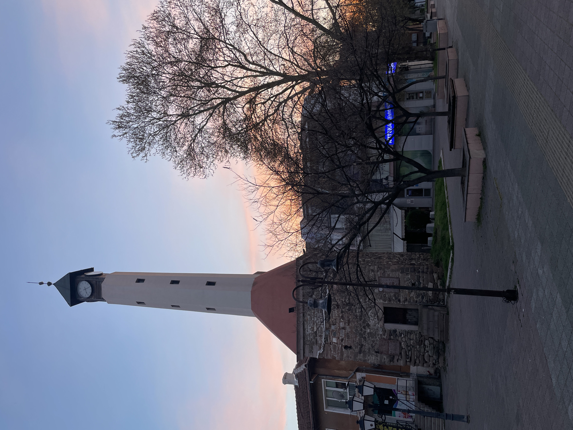 The image shows a street scene in Pazardzhik, Bulgaria, featuring the prominent Clock Tower (Sahat Tepe). The tower, with its stone base, painted shaft, and clock face, is positioned prominently against a backdrop of a pastel-colored sky, likely at sunrise or sunset. Bare tree branches frame the upper part of the image, while a street lamp stands in front of the tower's base. Buildings and structures are visible nearby, indicating the urban context of the scene. The ground is paved, and there are other decorative trees or planters located alongside the road.