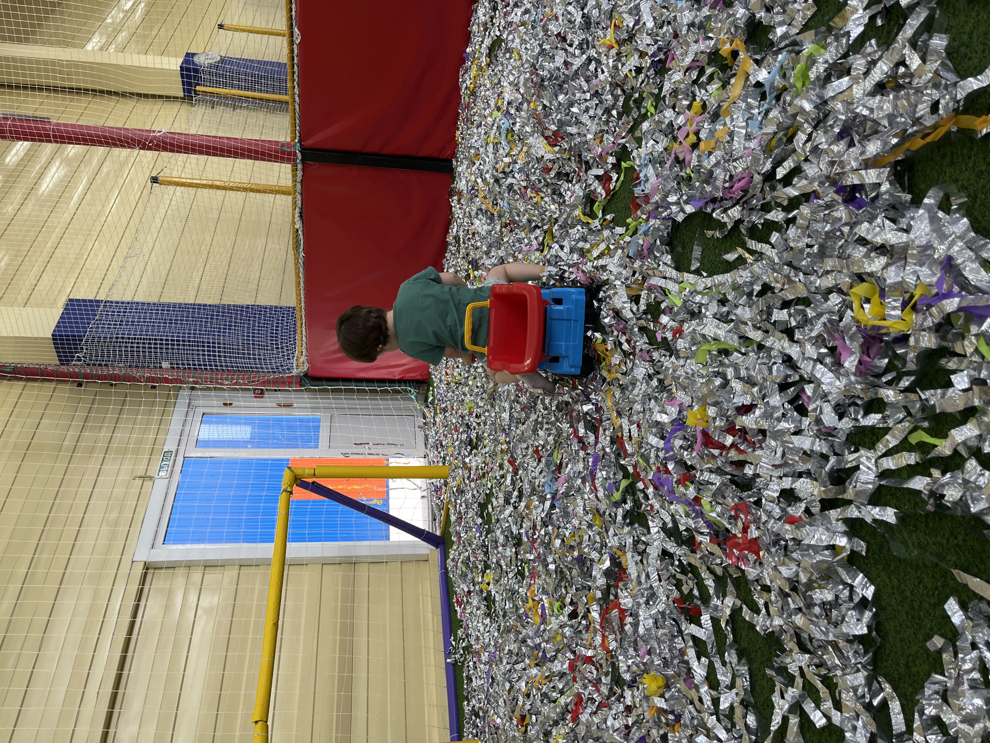 A child plays in a ball pit at a playground in Chaikovsky, Russia. The boy, wearing a green t-shirt, sits amongst shredded silver and multicolored confetti. He is holding a blue and red toy truck with a yellow handle. The ball pit floor appears to be green artificial turf. The scene is inside an indoor playground with padded walls, a window labeled "выход" (exit), and a netted enclosure.