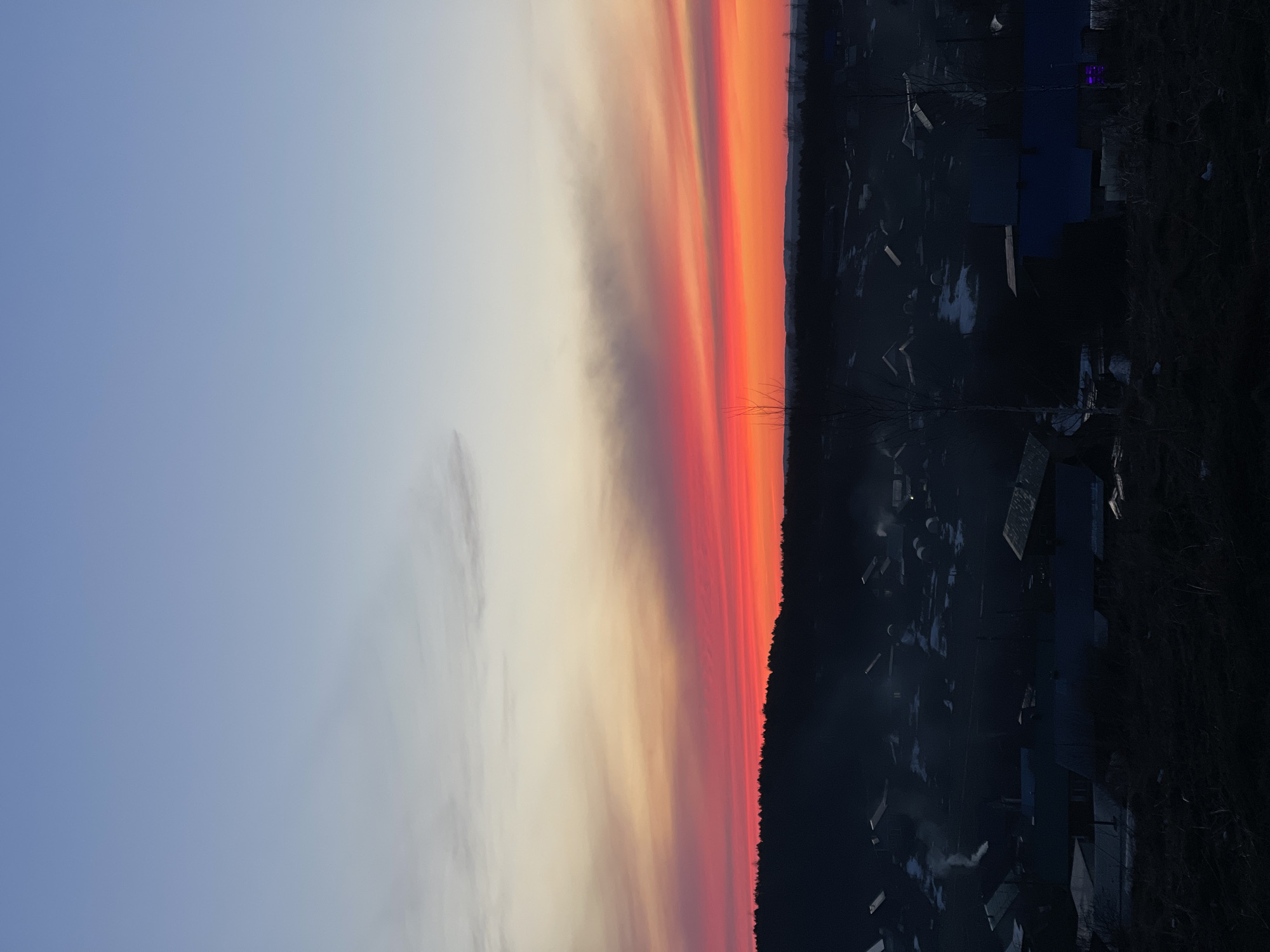 The image captures a sunset scene over Фоки, Russia. The sky transitions from light blue to shades of orange and red, with cloud formations visible. Below the horizon, a dark silhouette of a residential area with buildings and trees is present. There are no people visible in the image.