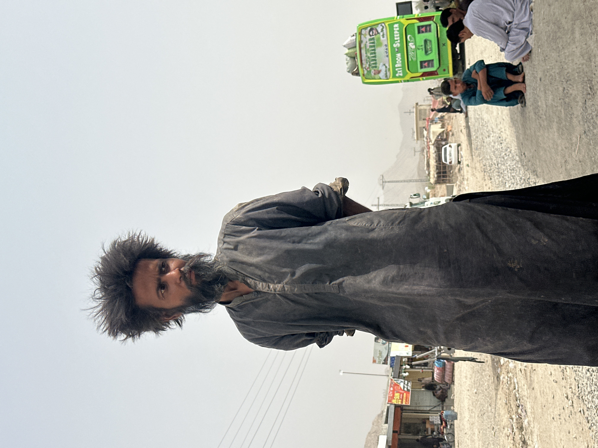 In the city of Khuzdar, Pakistan, a man with disheveled hair and a long beard stands in the foreground, looking to his right. He wears a dark, loose-fitting garment, possibly a shalwar kameez. In the background, a bright green bus with "SABARABAR" and "2x1 ROOM SLEEPER" written on it is parked on the side of a dusty road. Several other people are visible sitting on the ground near the bus, some in traditional clothing. The environment appears arid, with low-lying buildings and distant mountains under a hazy sky. The overall scene suggests a roadside stop or a gathering point along a travel route in a semi-urban or rural setting.