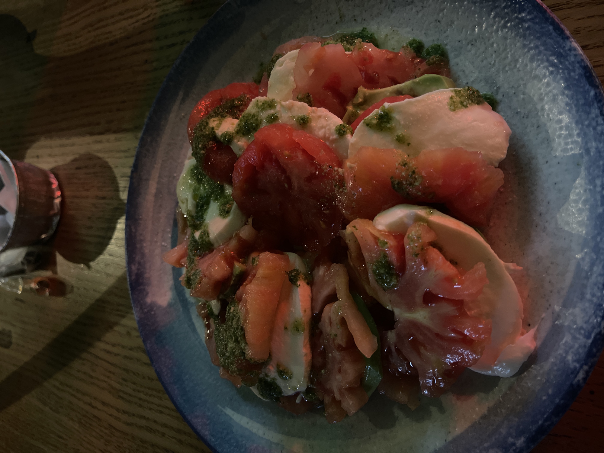 A close-up shot displays a caprese salad on a blue-rimmed plate, situated on a wooden table. The salad comprises sliced tomatoes, mozzarella cheese, and what appears to be pesto sauce drizzled on top. A small, metallic object sits on the table to the left of the plate. The image appears to be taken indoors under dim, warm lighting. There is no information to specify if the image was taken in Sofia, Bulgaria.