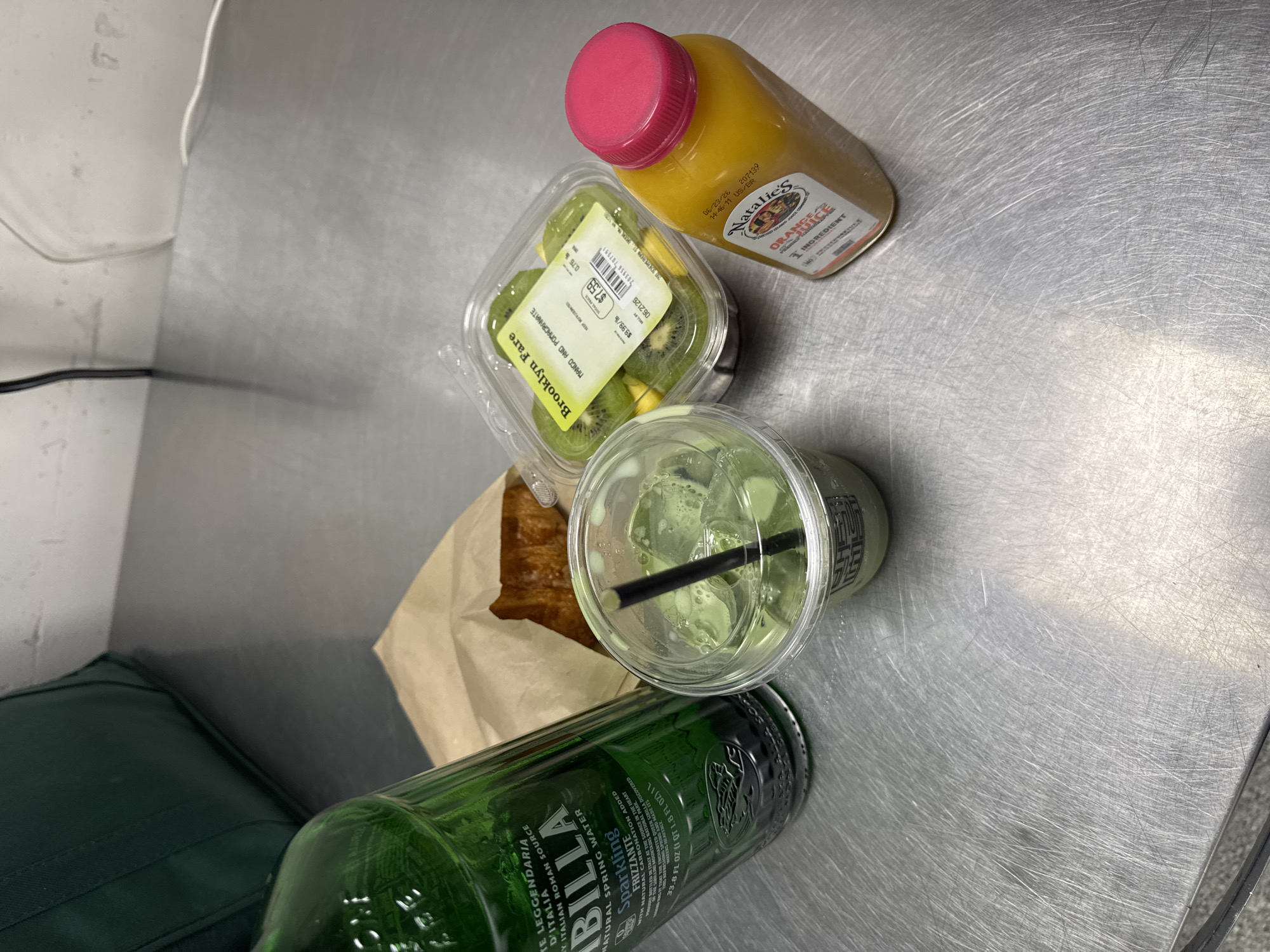A Brooklyn Fare branded plastic container filled with sliced kiwi and mango sits on a stainless steel counter. Next to it is a clear plastic cup with ice and a green beverage, likely matcha, with a black straw. A brown paper bag containing a pastry is behind the cup. To the left is a green glass bottle of San Pellegrino water. To the right is a bottle of Natalie's Orange Juice. The scene is indoors on a metal counter, likely in Brooklyn, New York.