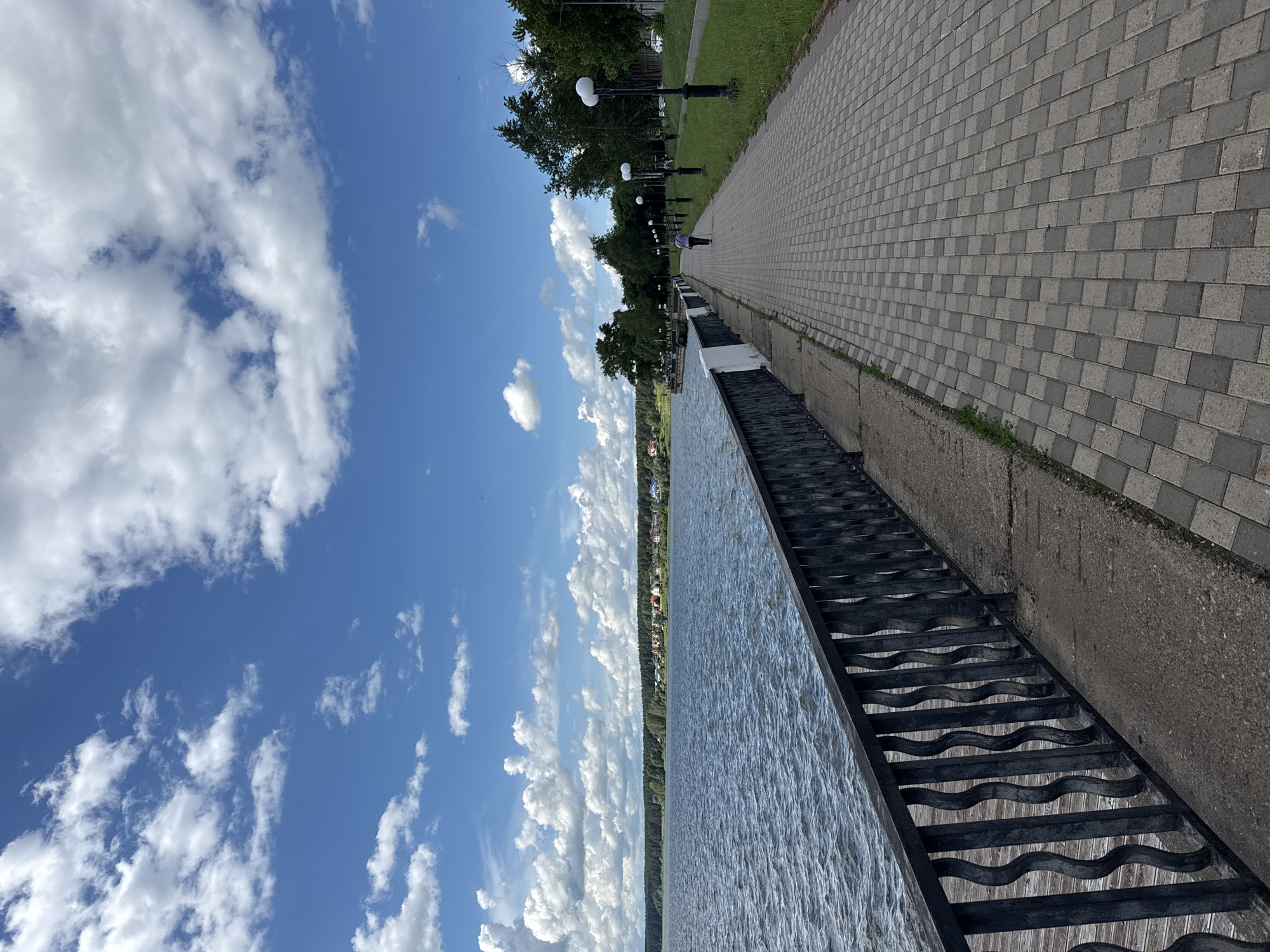 A paved walkway runs along the shore of a body of water, bordered by a black metal railing. The water is choppy, reflecting the blue sky and scattered white clouds. On the far side of the water, a line of trees and buildings is visible. The walkway is lined with lampposts and grass, with trees further back on the right side of the image. A single person is walking away from the camera on the walkway, wearing a purple shirt and dark pants. The scene appears to be a public park or promenade. The location is likely the embankment in Votkinsk, Russia, a city known for its picturesque waterfront and association with composer Pyotr Ilyich Tchaikovsky.