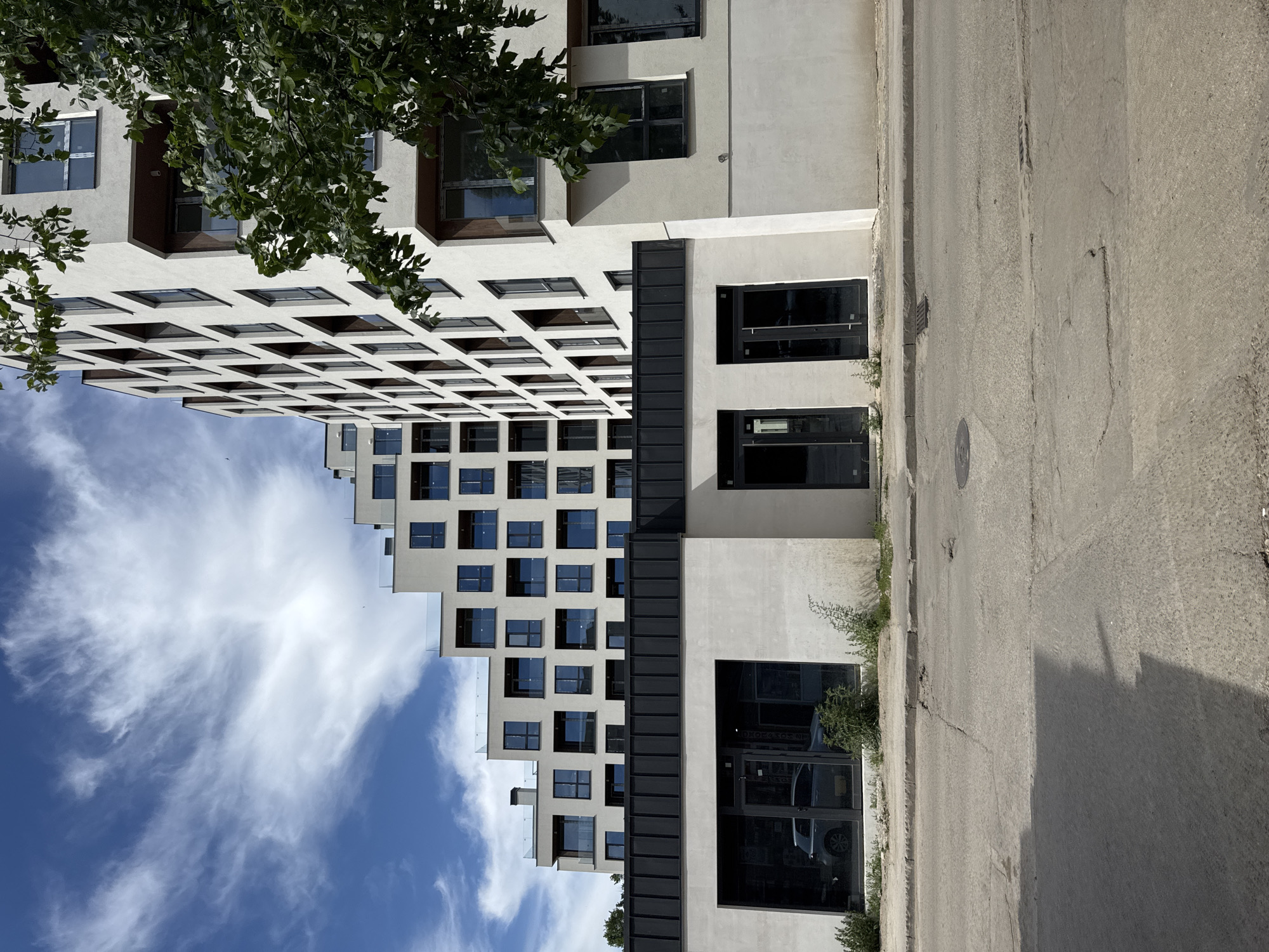 The image depicts a view in Sofia, Bulgaria, capturing the architecture of two adjacent buildings and a street. The first building is a modern high-rise, characterized by a stepped design with numerous square windows against a light facade, partially obscured by tree foliage at the top. The sky, with scattered clouds, provides a contrasting backdrop. The second building is lower, features a light-colored facade punctuated by dark-framed glass openings. In front of the buildings, there is a paved road surface.